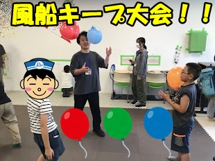 Instructor guiding children in a balloon game indoors, with a yellow banner overhead reading '風船キーフ大会!!'.