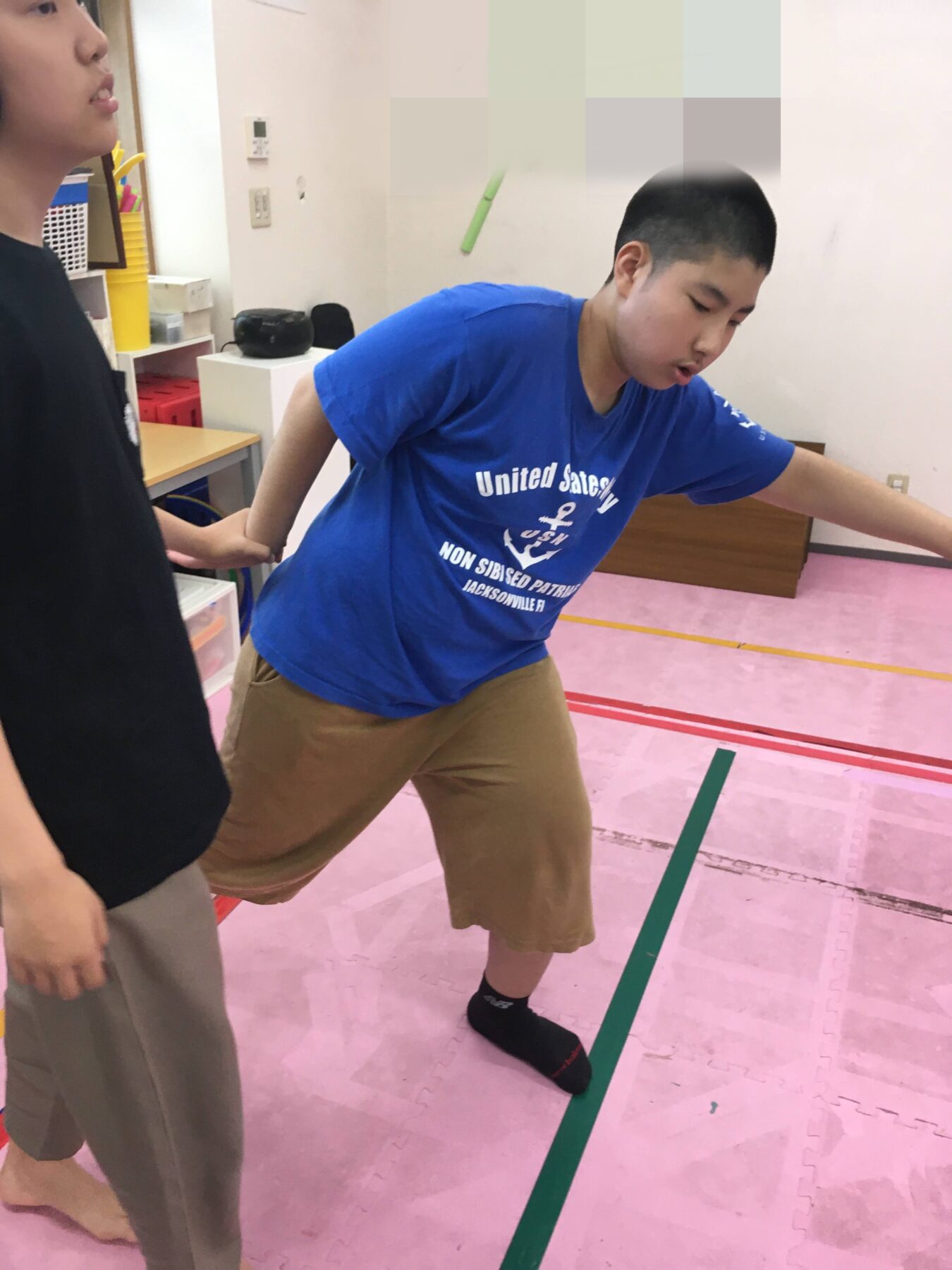Boy in a blue t-shirt leans forward while someone holds his left arm behind him in a room with pink foam mats.