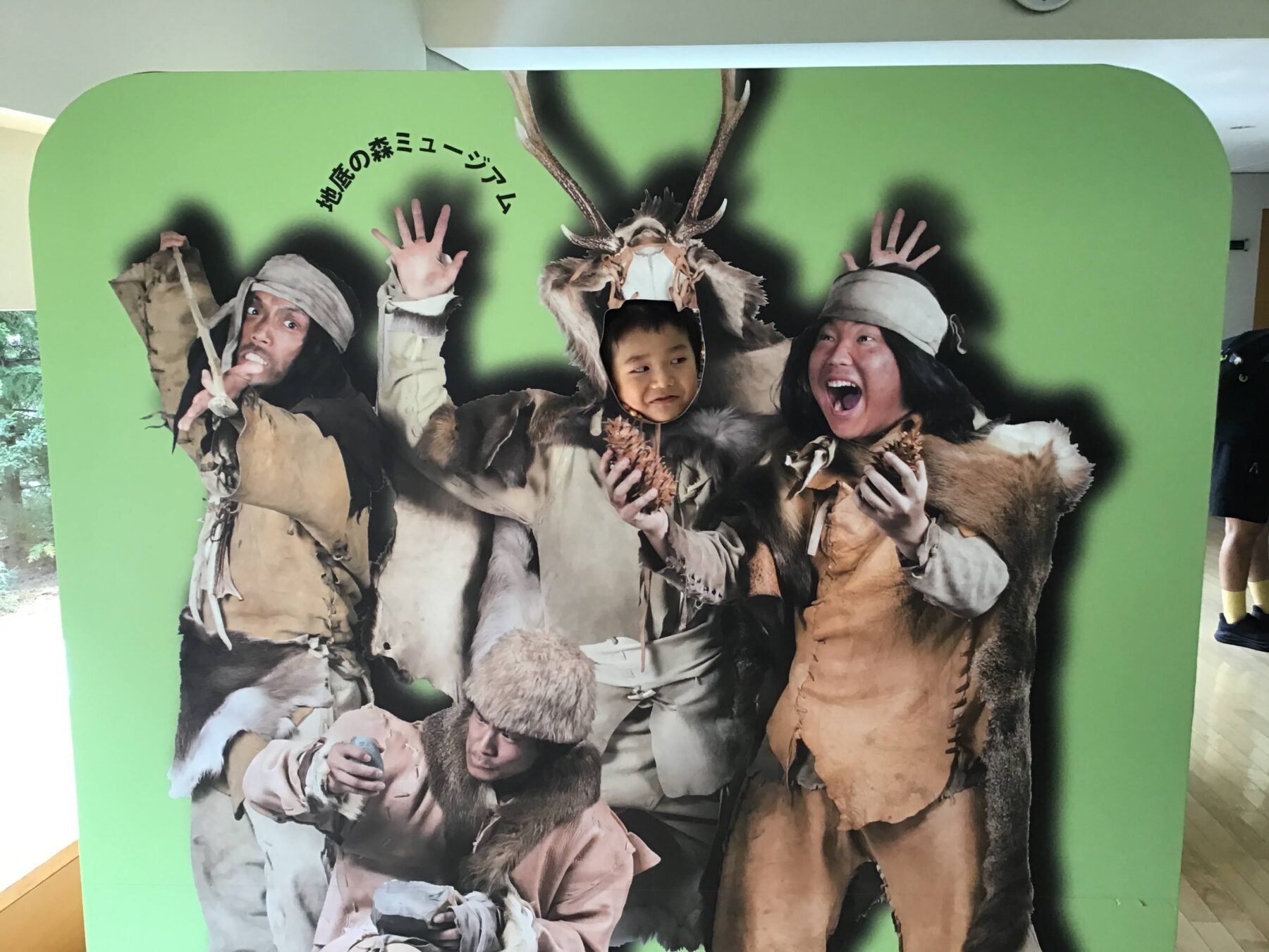 Child sticks head through a life-size photo cutout of people in fur costumes posing with antlers, green backdrop behind them (fun museum display)