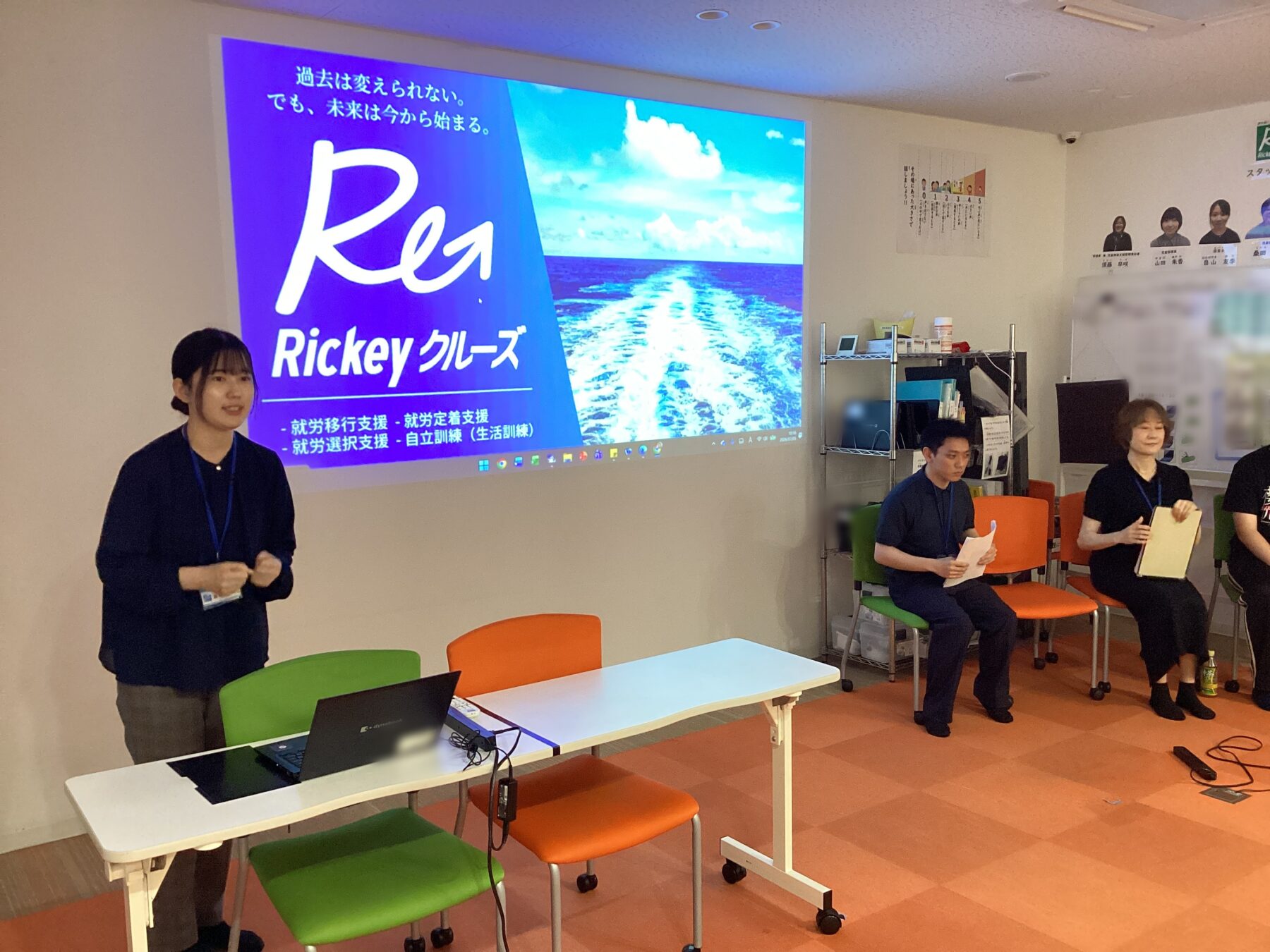 Presenter standing near a projection screen with a 'Ricky Cruise' slide and ocean image, addressing a small group in a classroom.