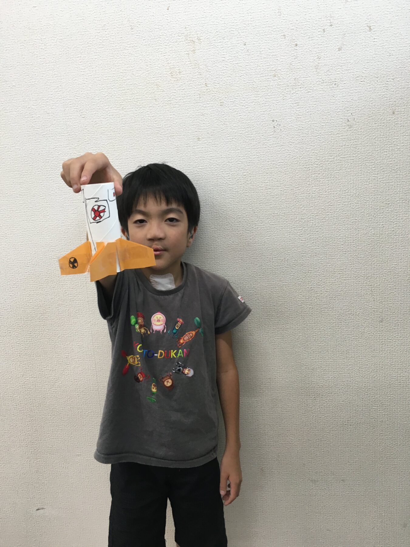 A boy holding up a white paper craft with orange origami fins against a plain wall.