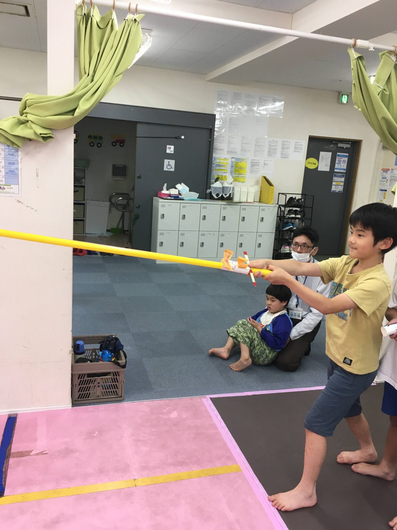 Boy in a yellow shirt holds a long yellow stick during a group activity in a gym, with a seated child and an adult nearby.