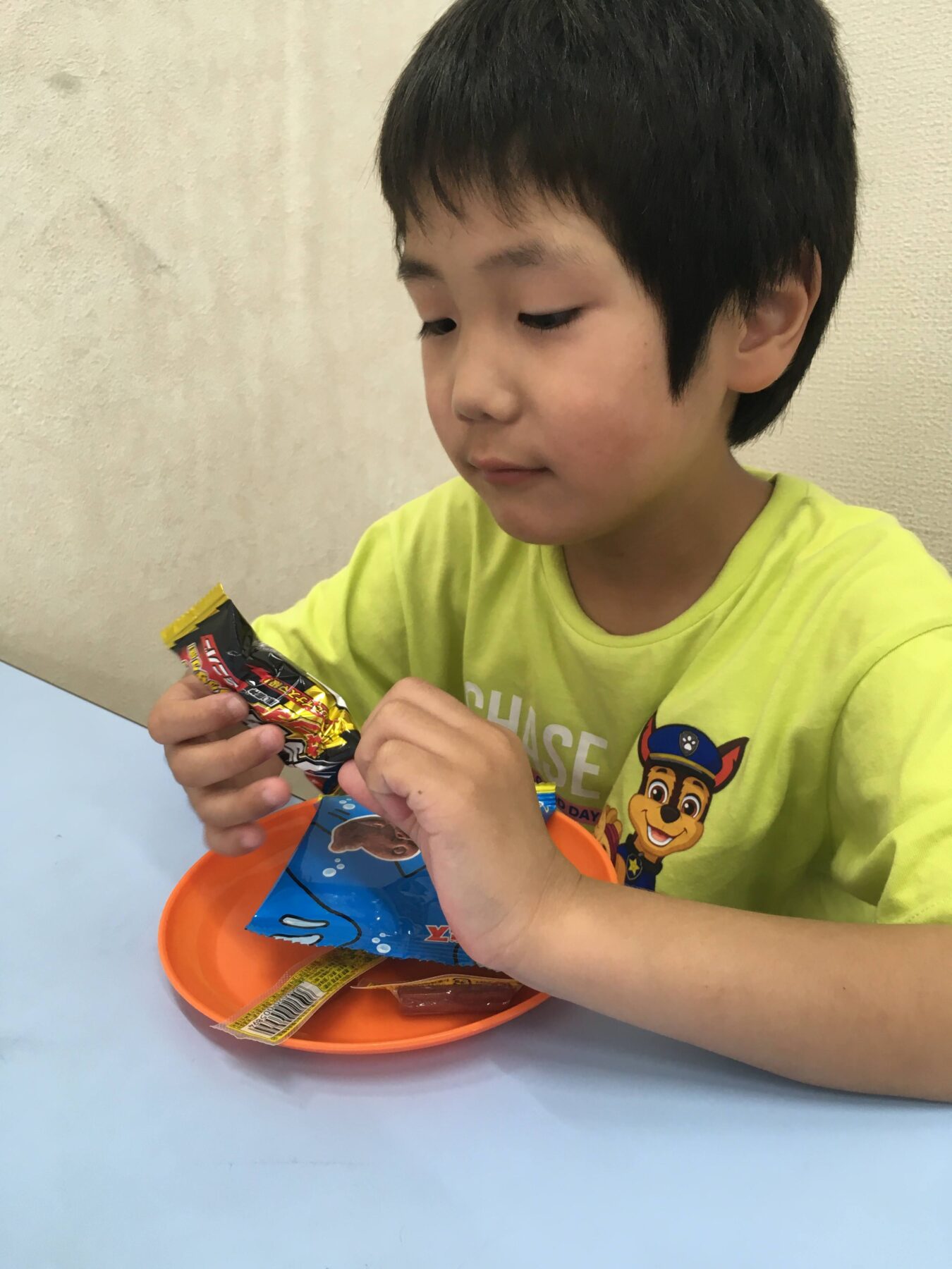 Young boy in a lime shirt unwraps a candy bar over an orange bowl on a light blue table.