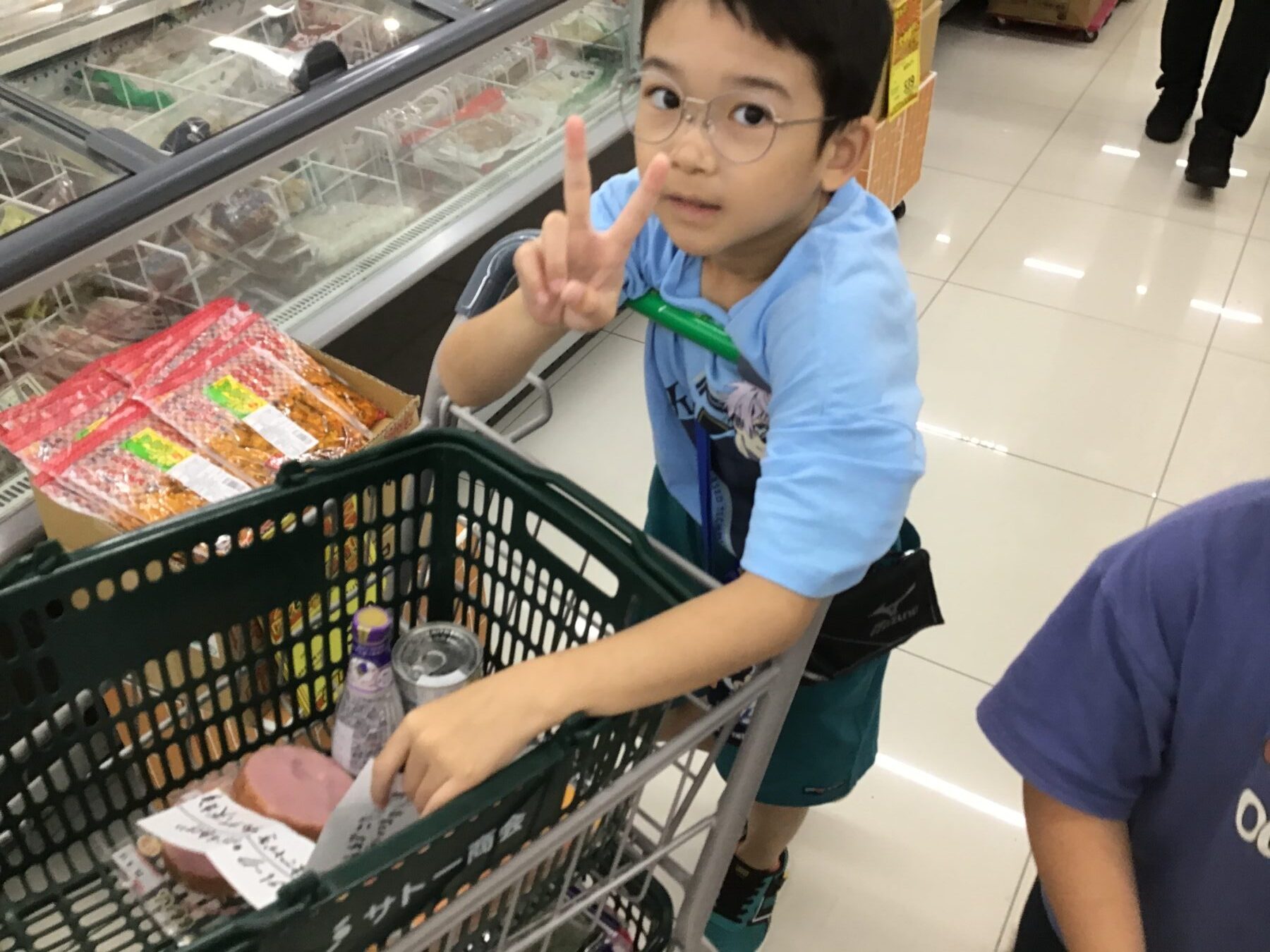 Child in a store cart making a peace sign, blue shirt and glasses, groceries in the cart nearby.