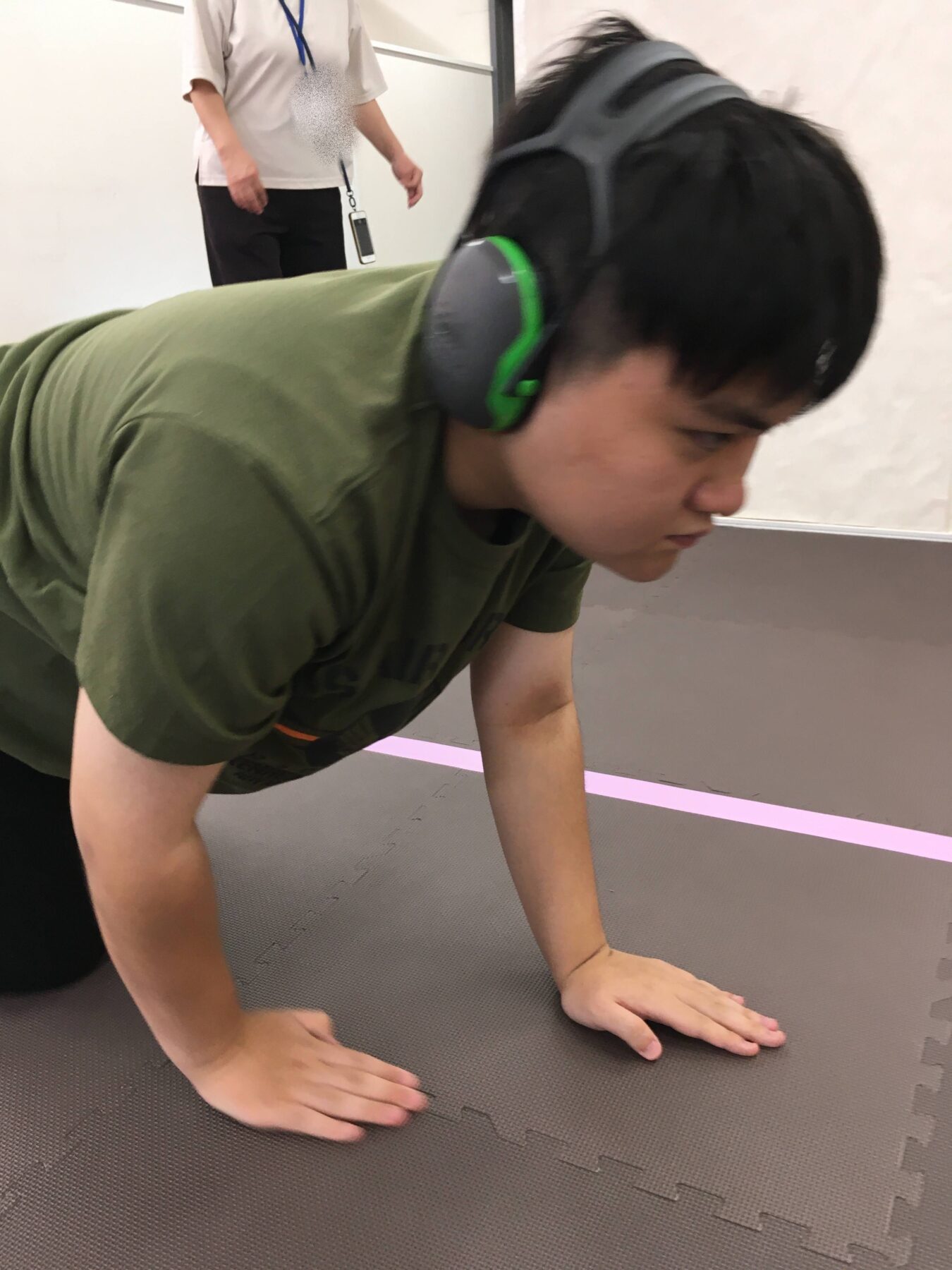 Person on hands and knees in a green shirt doing a plank-like exercise on a mat, wearing large green and gray headphones.