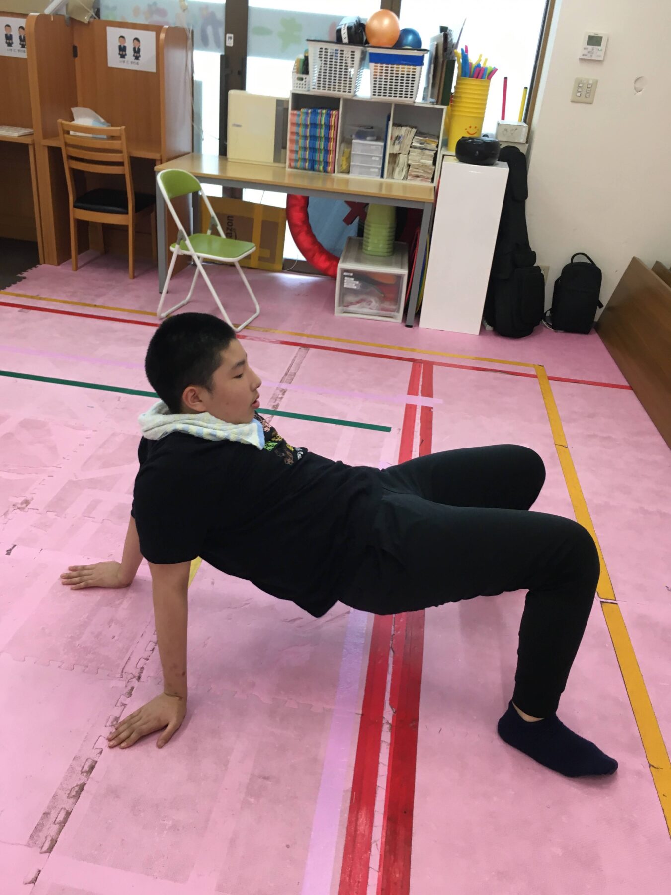 Child doing a low plank exercise on a pink gym floor in a classroom/playroom setting, supporting weight on hands and toes with a towel around neck/shoulder area visible