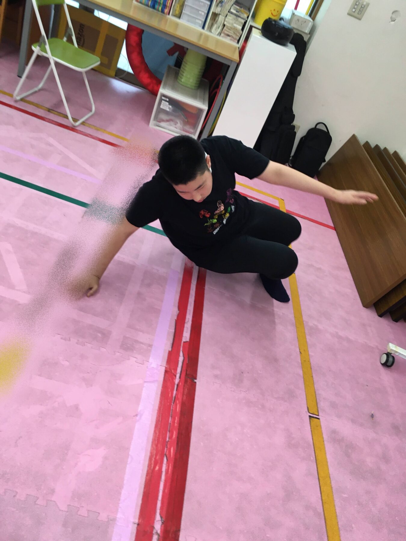 Boy in a black shirt crouches on a pink padded floor with colorful lines, balancing with arms outstretched in a playroom.