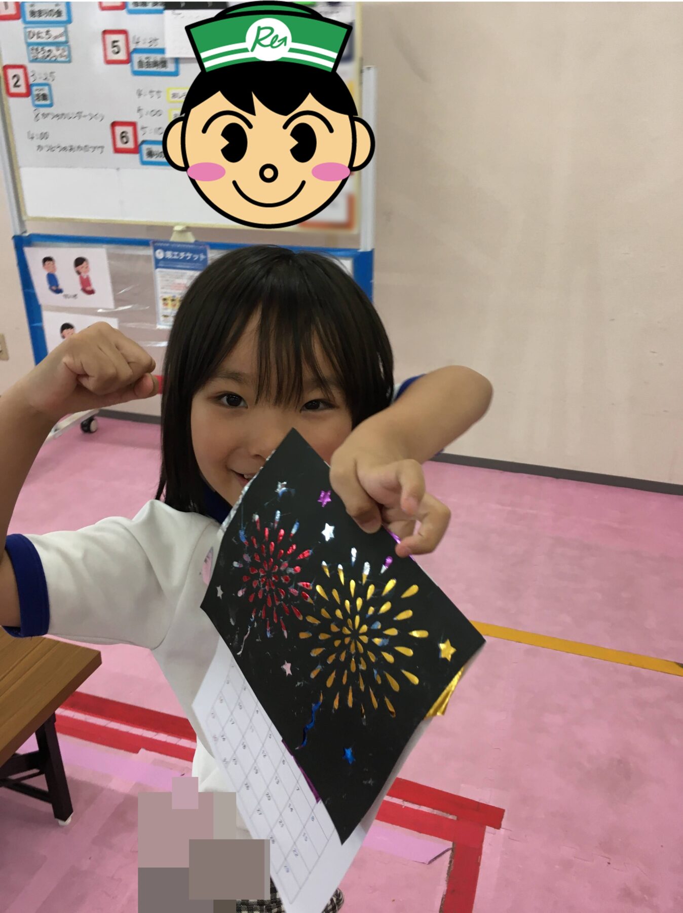 Young girl in a white shirt with blue trim holds up a black card featuring colorful fireworks, smiling and pointing at it in a classroom.