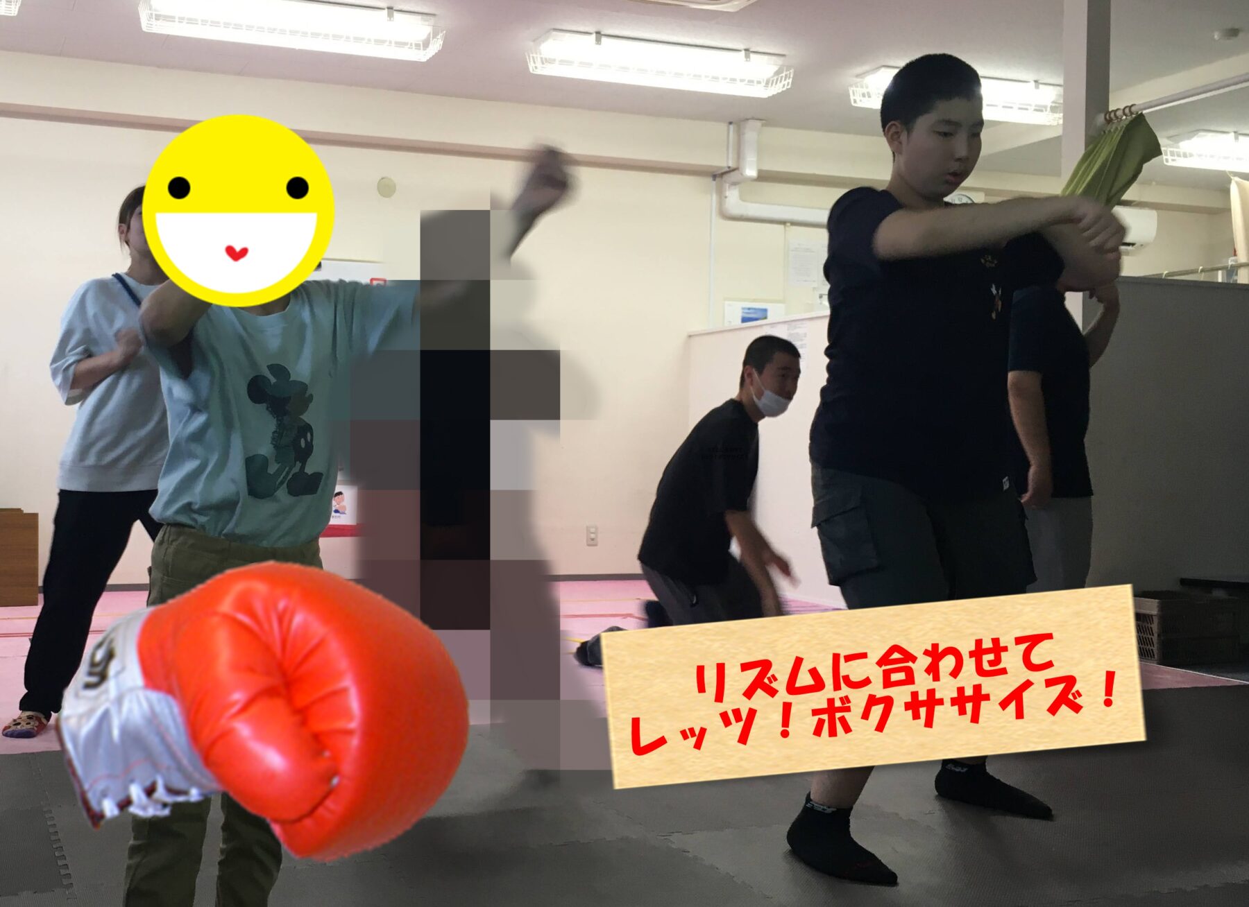 In a gym or martial arts room, a person in a light blue shirt throws a punch toward the camera, with a large orange boxing glove visible in the foreground; other students stand in the background, one wearing a mask. A yellow smiley emoji covers the front person's face. A wooden sign with red Japanese text is at the bottom right.