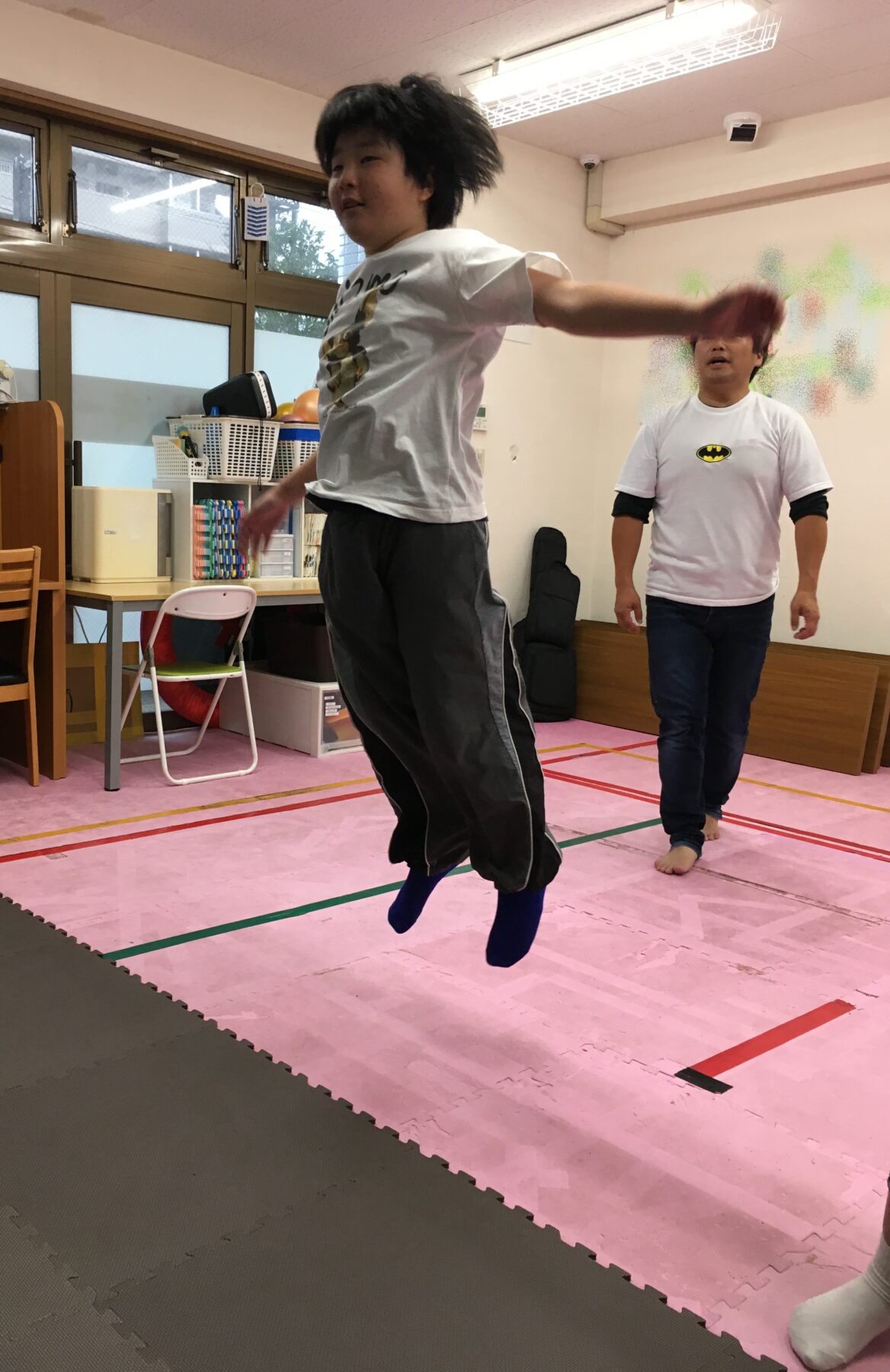 Person mid-jump with arms extended on pink foam mats; another person watches in a room with gym gear in the background.