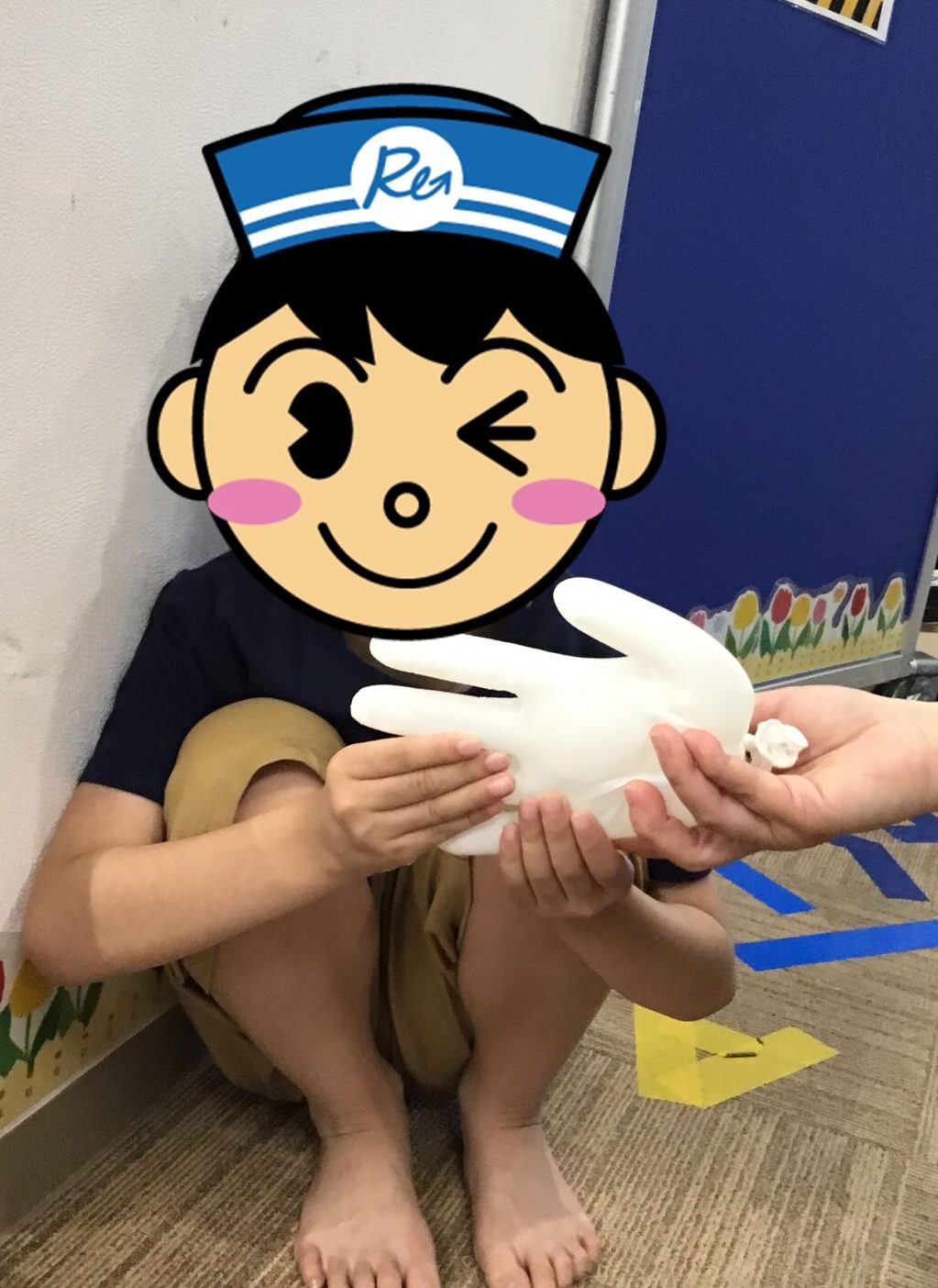 Child sitting barefoot on the classroom carpet, holding a large white glove prop handed by an adult. A cartoon face sticker covers the child.