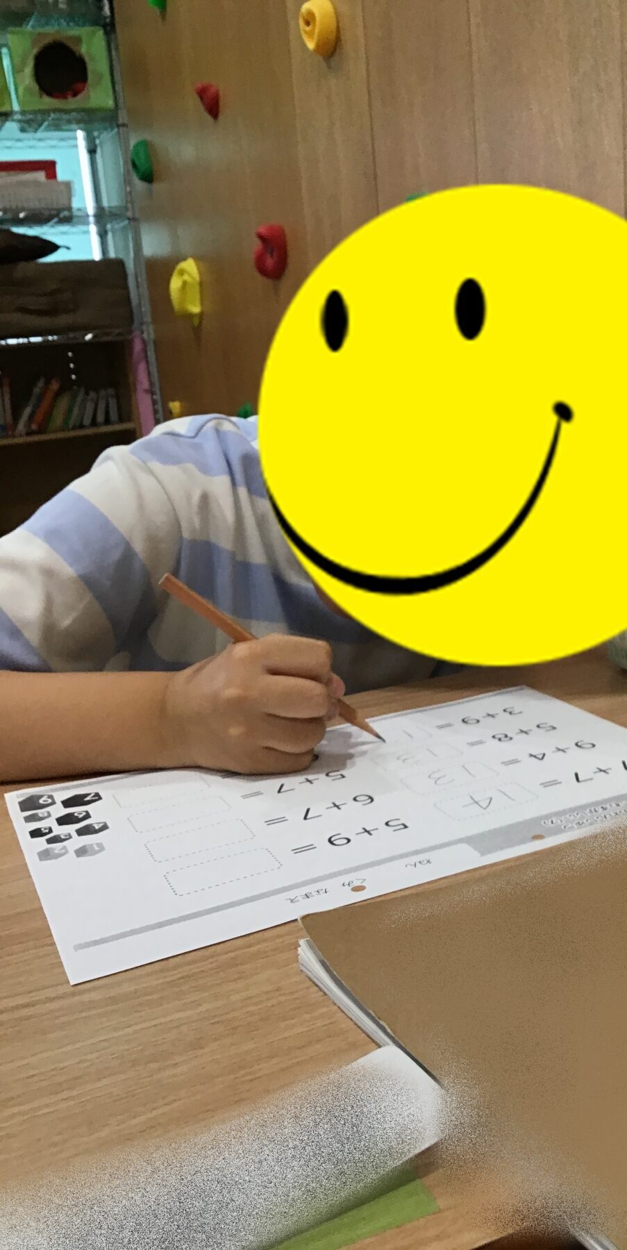 Child seated at a desk writing on a math worksheet with a pencil; a large yellow smiley overlay covers their face in a classroom setting.