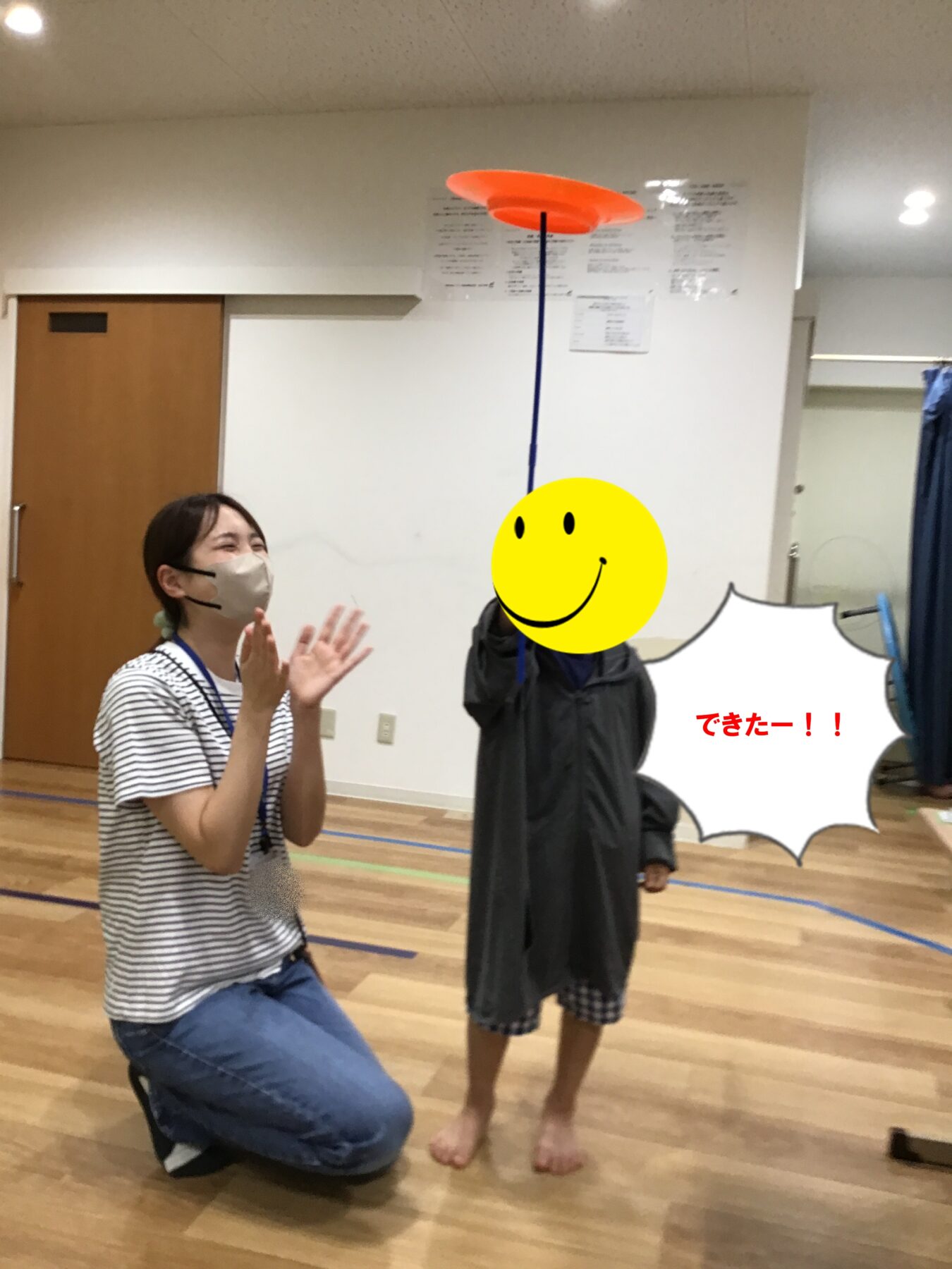 Woman in a striped shirt kneels and claps as a child balances a tall blue pole with an orange disk on top; speech bubble reads 'できたー!!'.