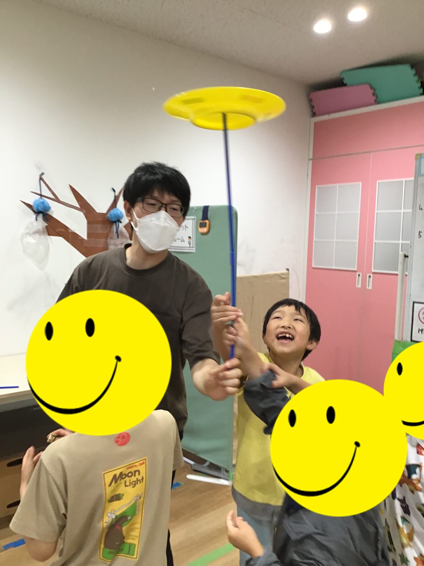 Person wearing a mask and glasses spins a yellow spinning disc on a long blue pole while children laugh in a colorful playroom.