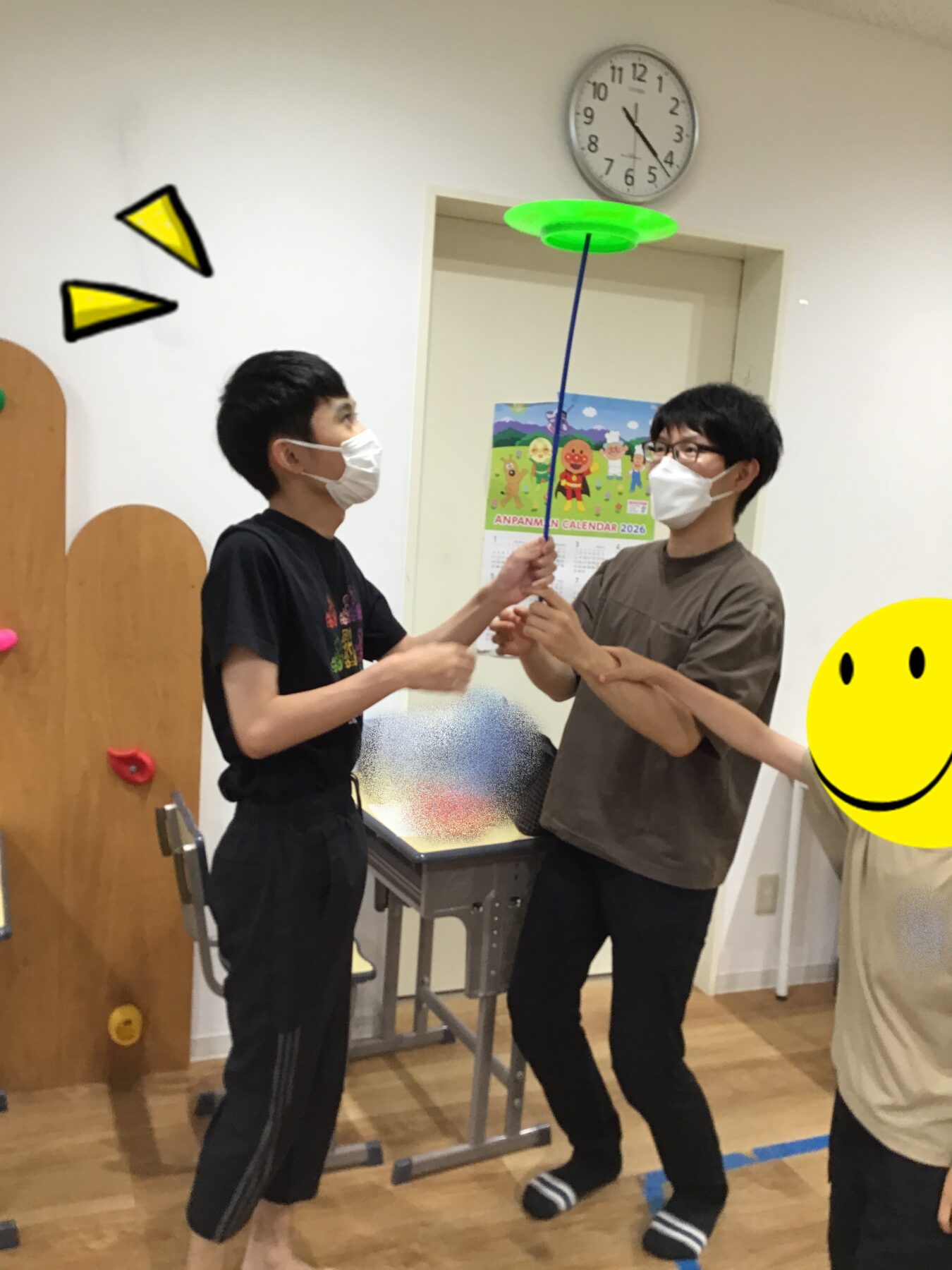 Two people wearing masks stand facing each other, each holding a blue pole with a bright green disc on top, indoors.