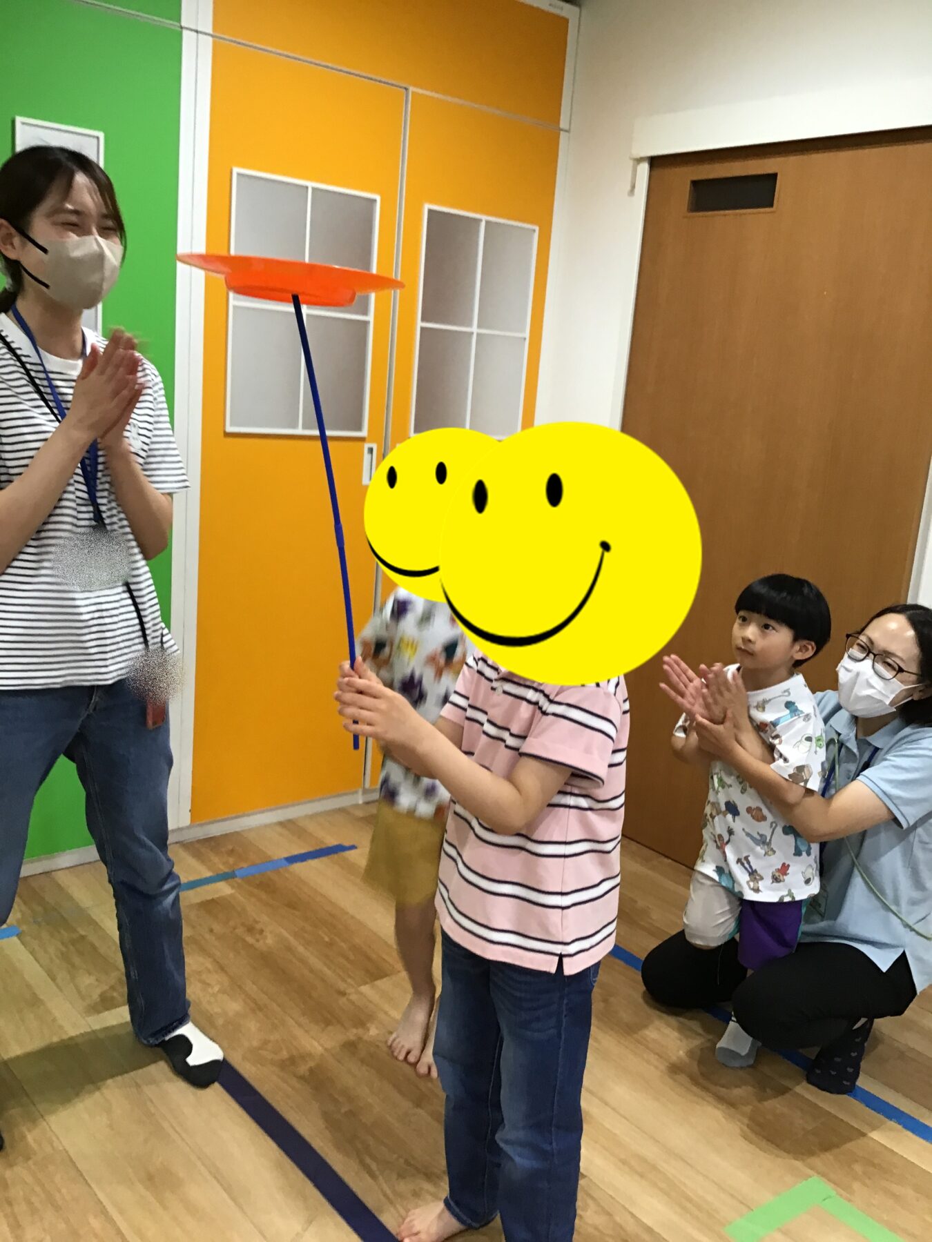 Teacher guides children in a play activity as a child spins an orange plate on a blue rod in a colorful room; all wear masks and watch eagerly.
