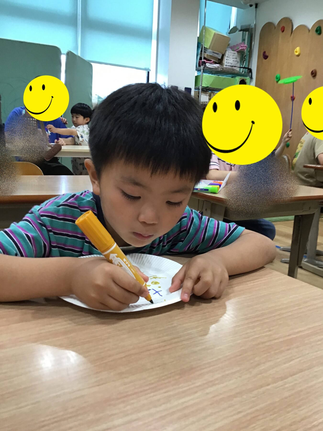 A young boy in a striped shirt sits at a wooden table drawing with an orange marker on a white paper plate-shaped sheet in a classroom setting.