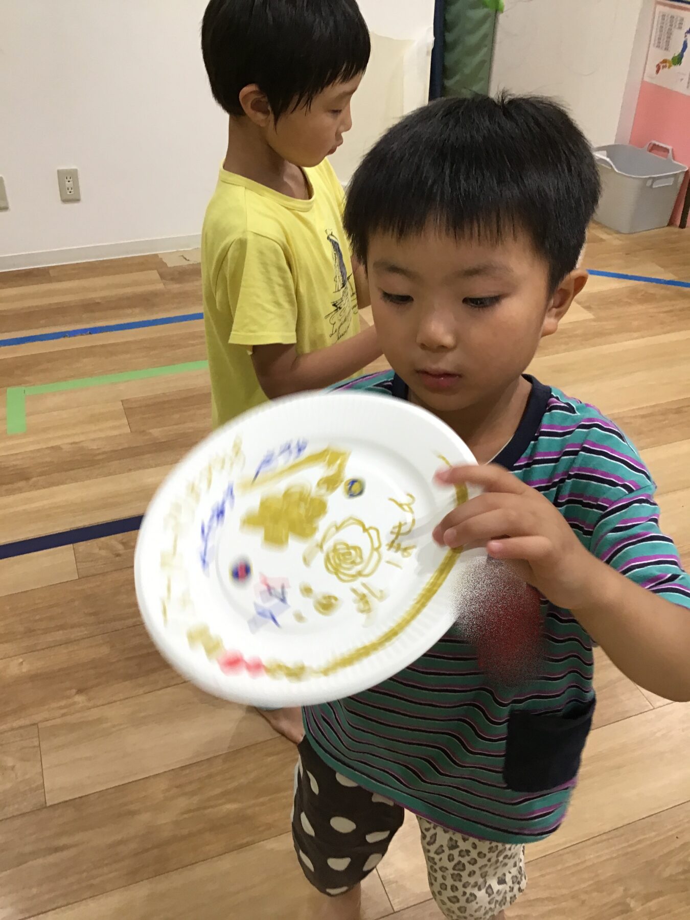 A young boy in a striped shirt holds up a decorated paper plate during an arts-and-crafts activity indoors.