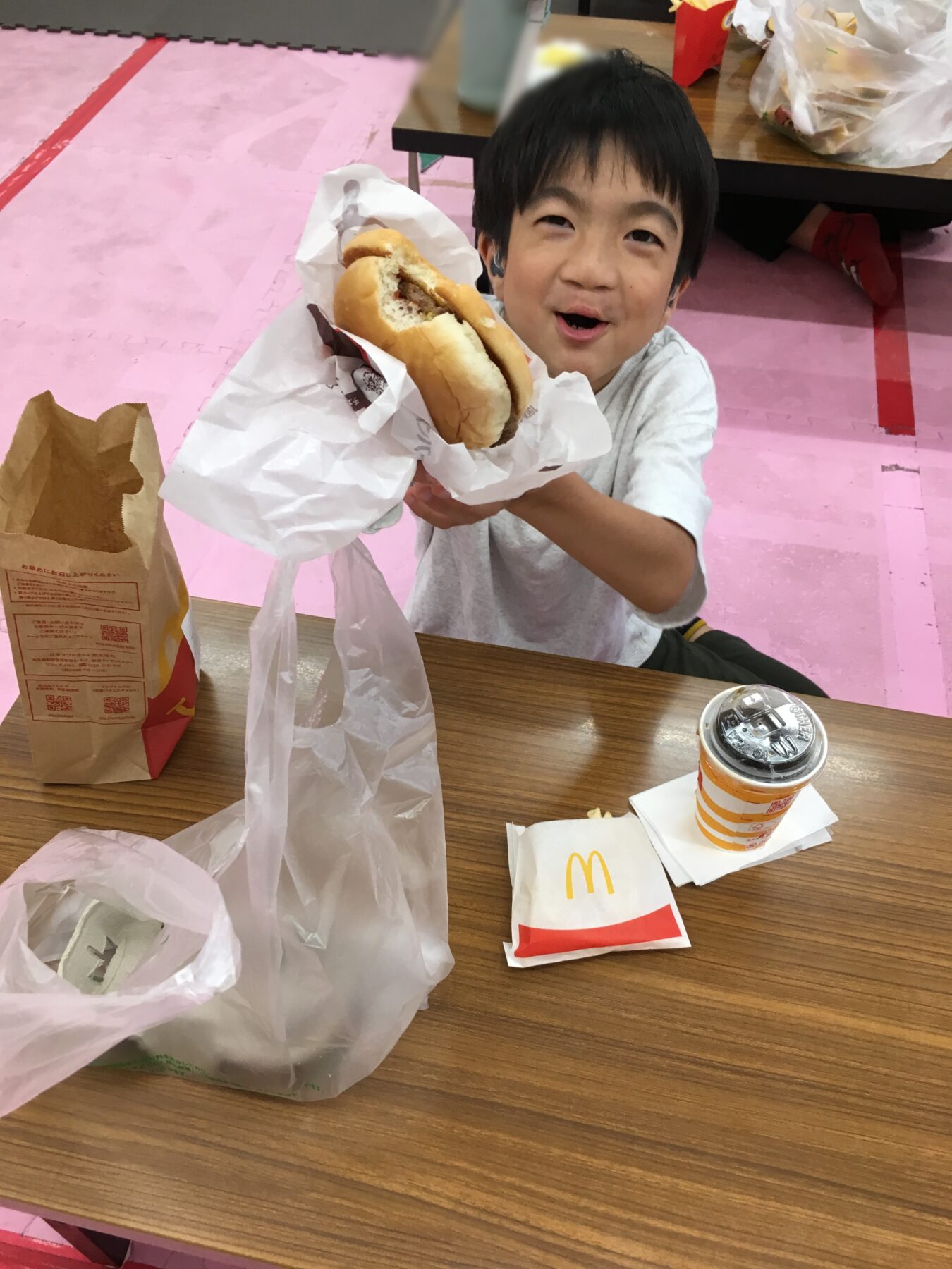 Young boy smiling and presenting a hamburger wrapped in paper at a McDonald's table.