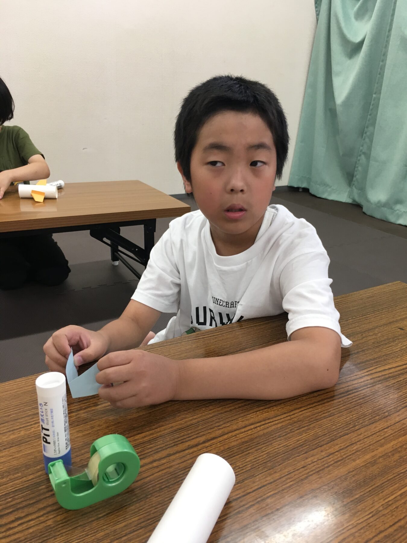 Child seated at a table making a craft with blue paper, glue stick, and tape on a wooden tabletop.