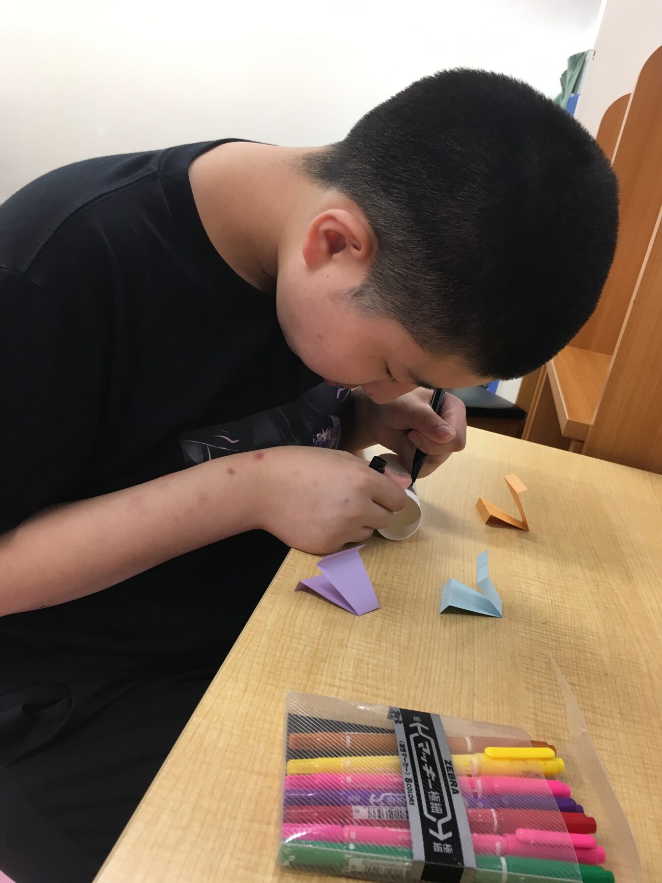 Child seated at a desk folding colorful origami pieces with markers and folded paper scraps nearby, focusing on crafts.