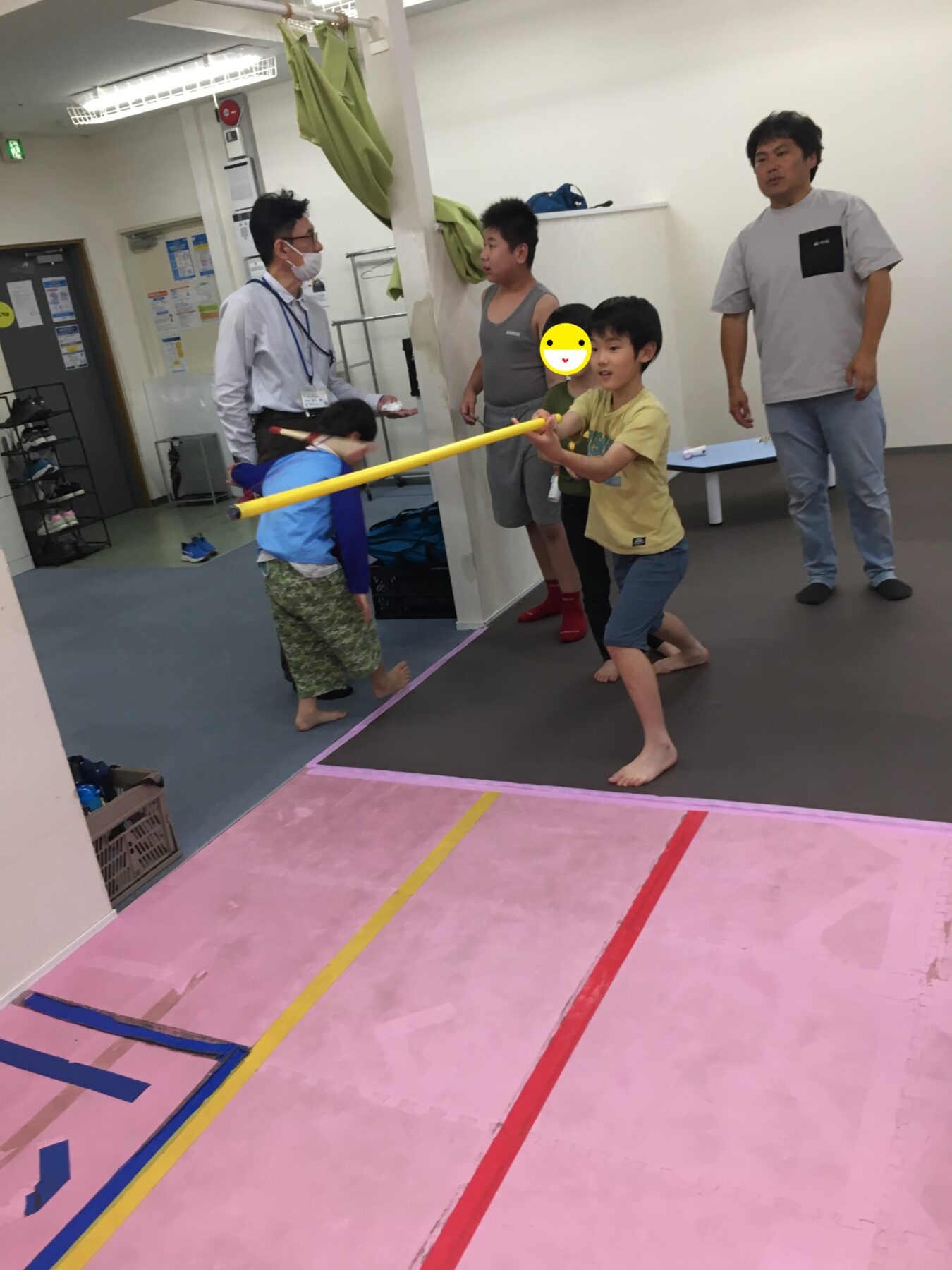 Children play with a yellow foam baton in a gym-like room, supervised by adults standing nearby.