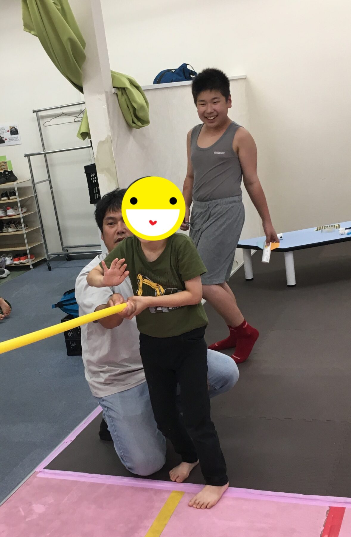 Child standing on a pink gym mat, holding a yellow pole with help from an adult kneeling beside them; both indoors in a gym.