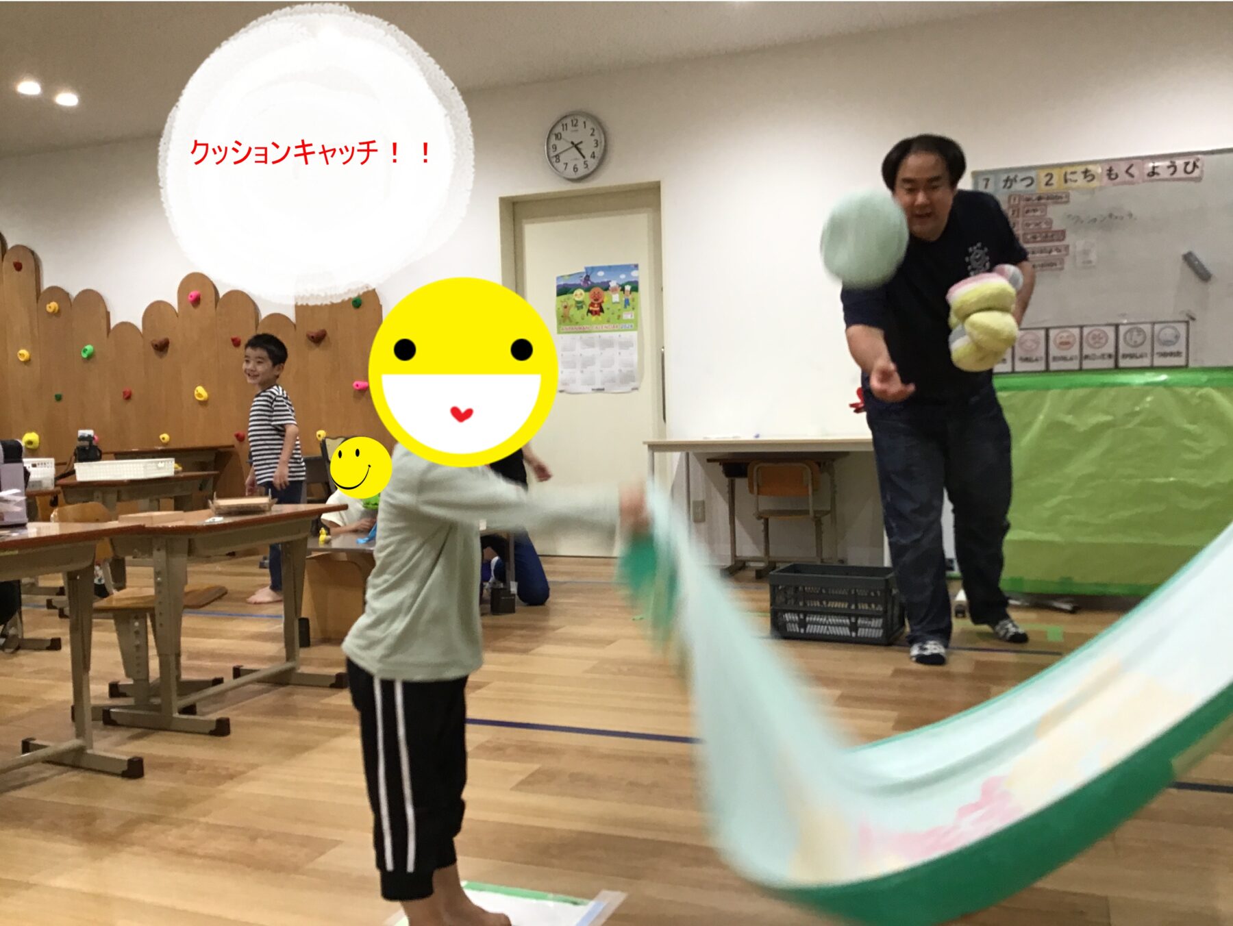 Cushion catch activity in a classroom gym; a man throws a pastel cushion to children, with others watching, and the word 'クッションキャッチ！！' shown on screen.