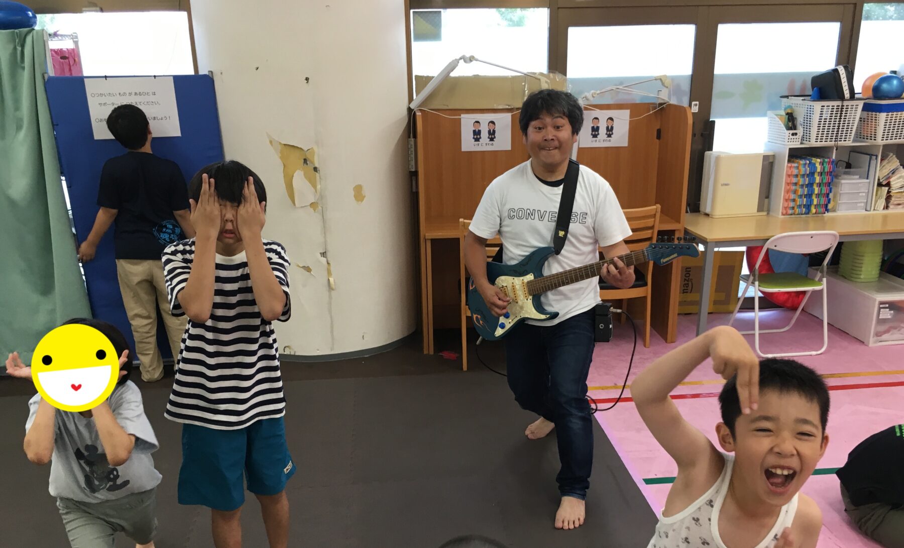 A man plays an electric guitar while smiling kids dance in a colorful classroom or playroom in the background.