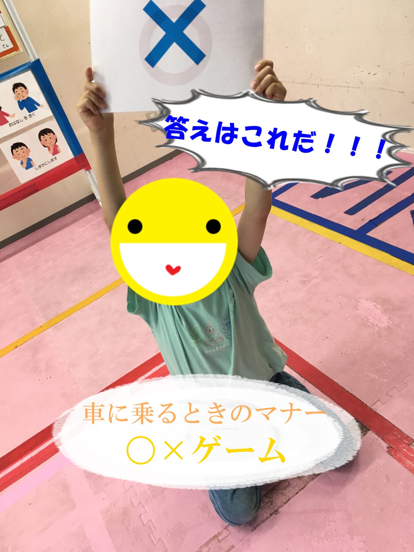 Child kneels on a pink gym floor, holding a paper with a blue X above, while a large speech-bubble-style caption in Japanese reads '答えはこれだ!!!' and a banner at the bottom mentions '車に乗るときのマナー OXゲーム' (an O/X game about car manners); the child's face is covered with a smiley emoji.