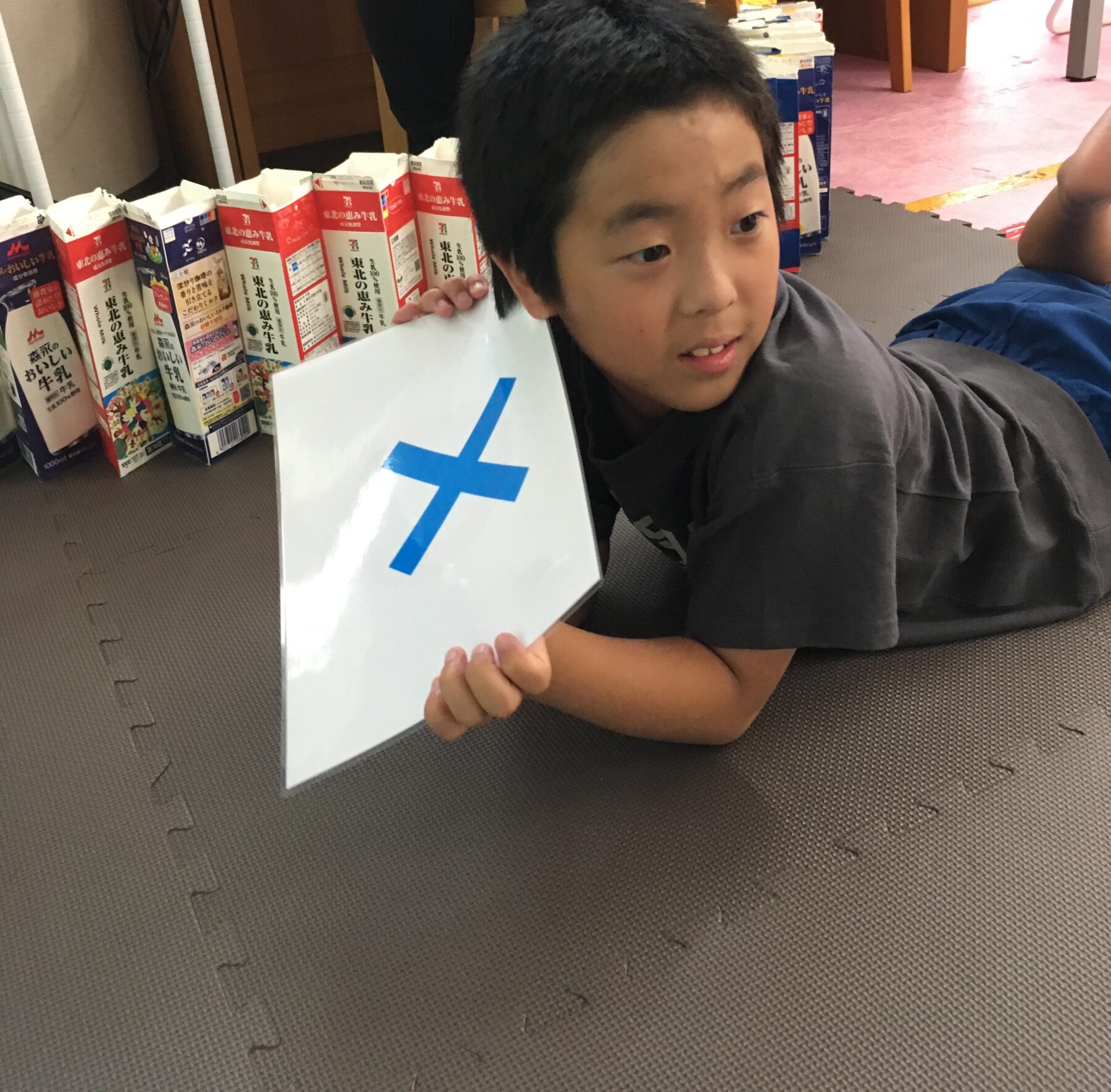 A young boy lying on a foam mat, propped on his side, holding a white sign with a blue cross, with boxed drinks lined up behind him.