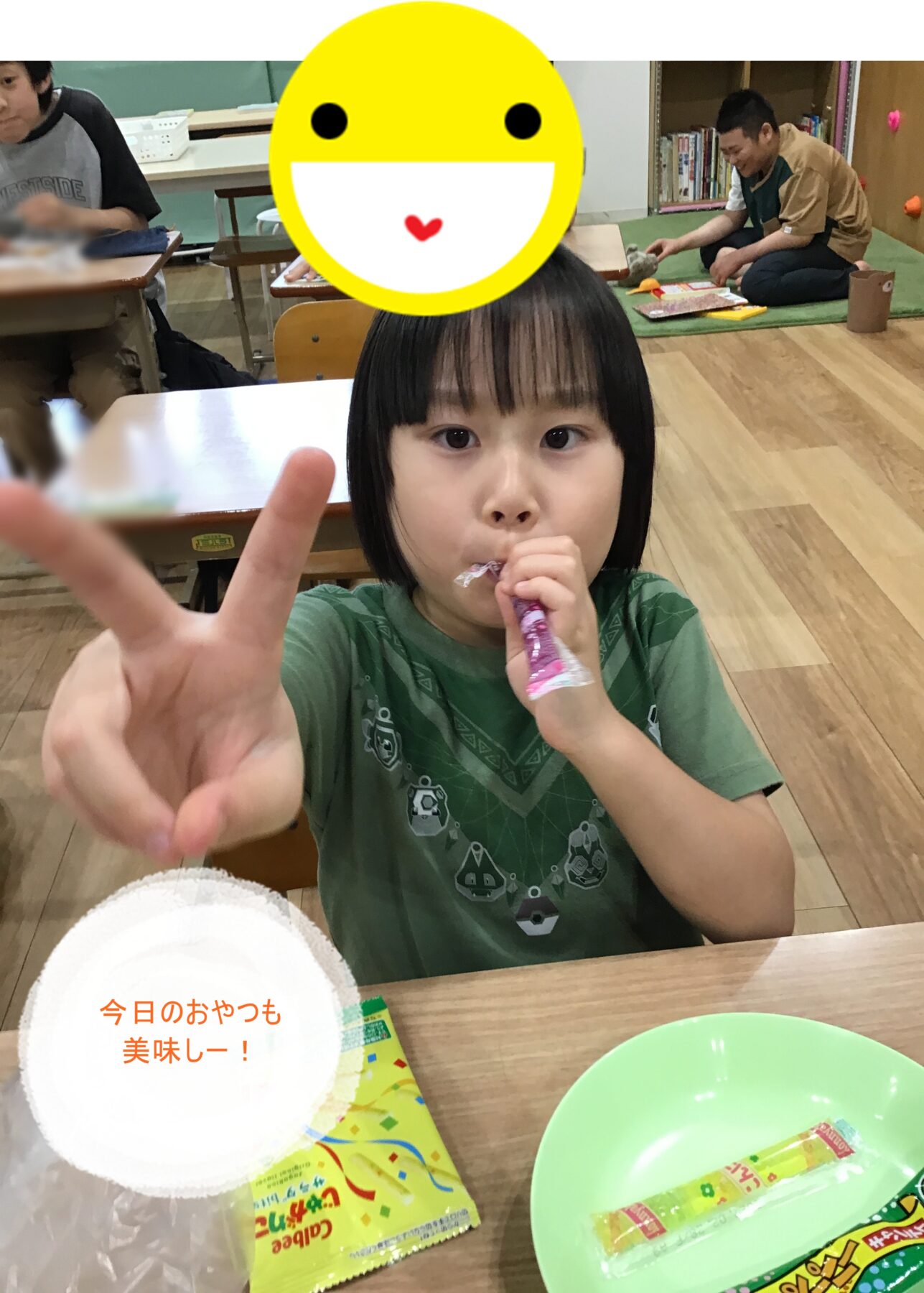 Child at a table posing with a peace sign while eating a wrapped snack; a large yellow smiley sticker covers their face in the foreground.