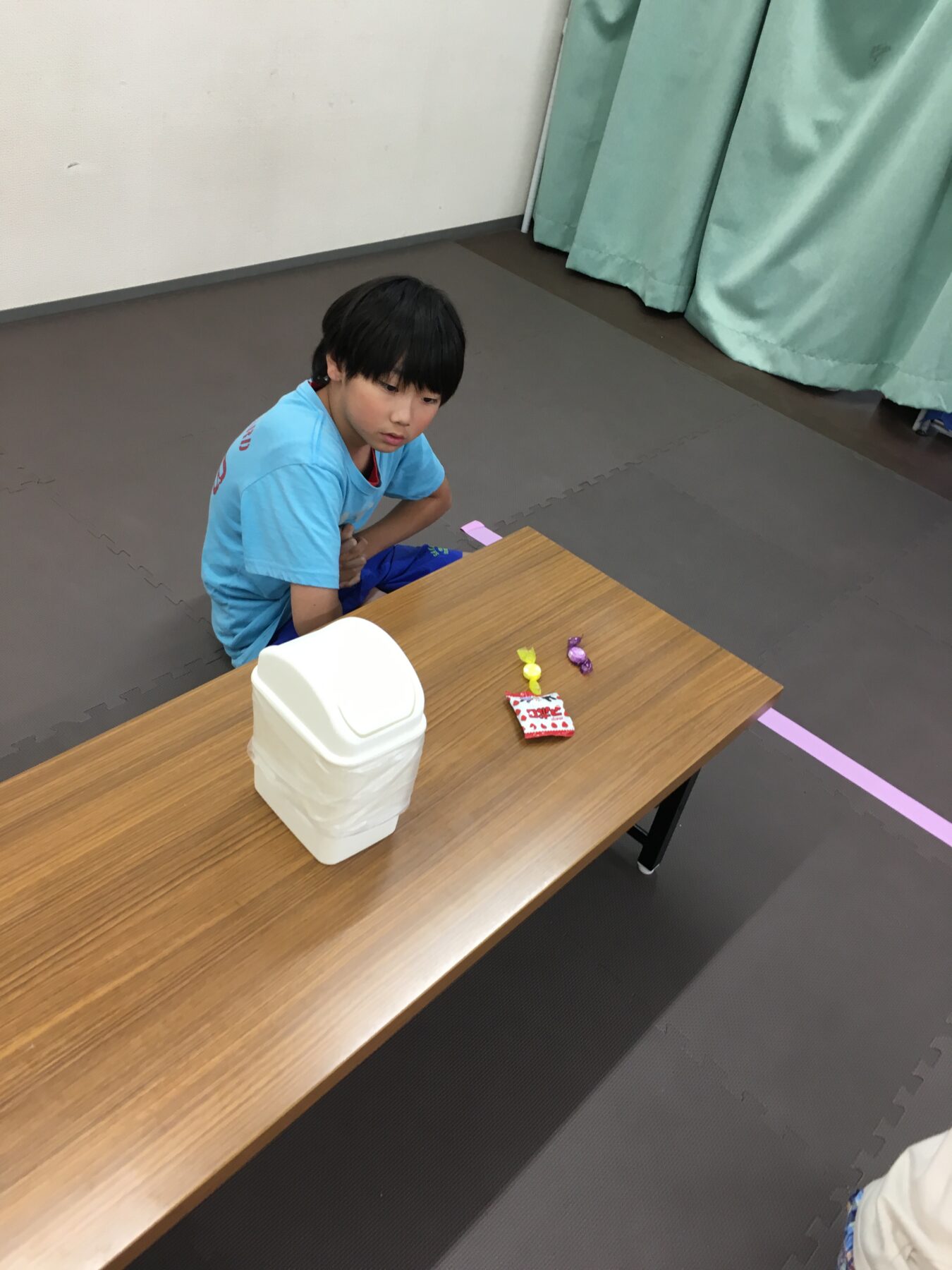 Young boy in a blue shirt sits at a wooden table, looking at colorful wrapped candies and a white plastic container on a gym-style floor.