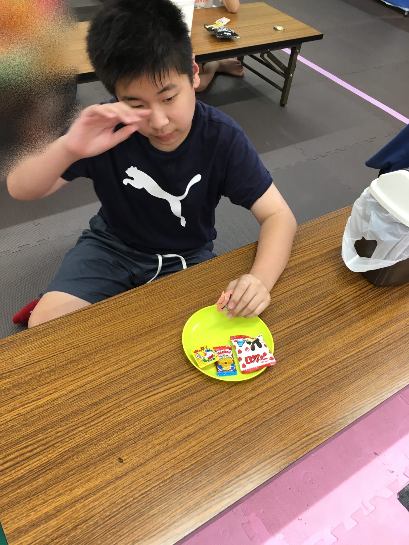 Young person in a navy Puma shirt sits at a wooden table, wiping their eye; a yellow plate with snack packets is in front.
