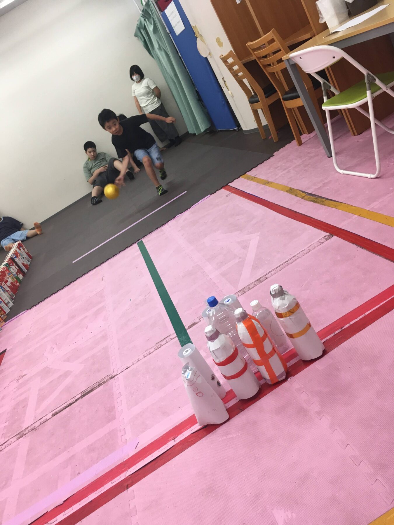Three children playing inside on pink foam mat, one rolling a yellow ball toward a row of bowling pins; others watch nearby.