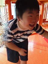 Smiling young child in a striped shirt and dark shorts, bending forward indoors on an orange floor.