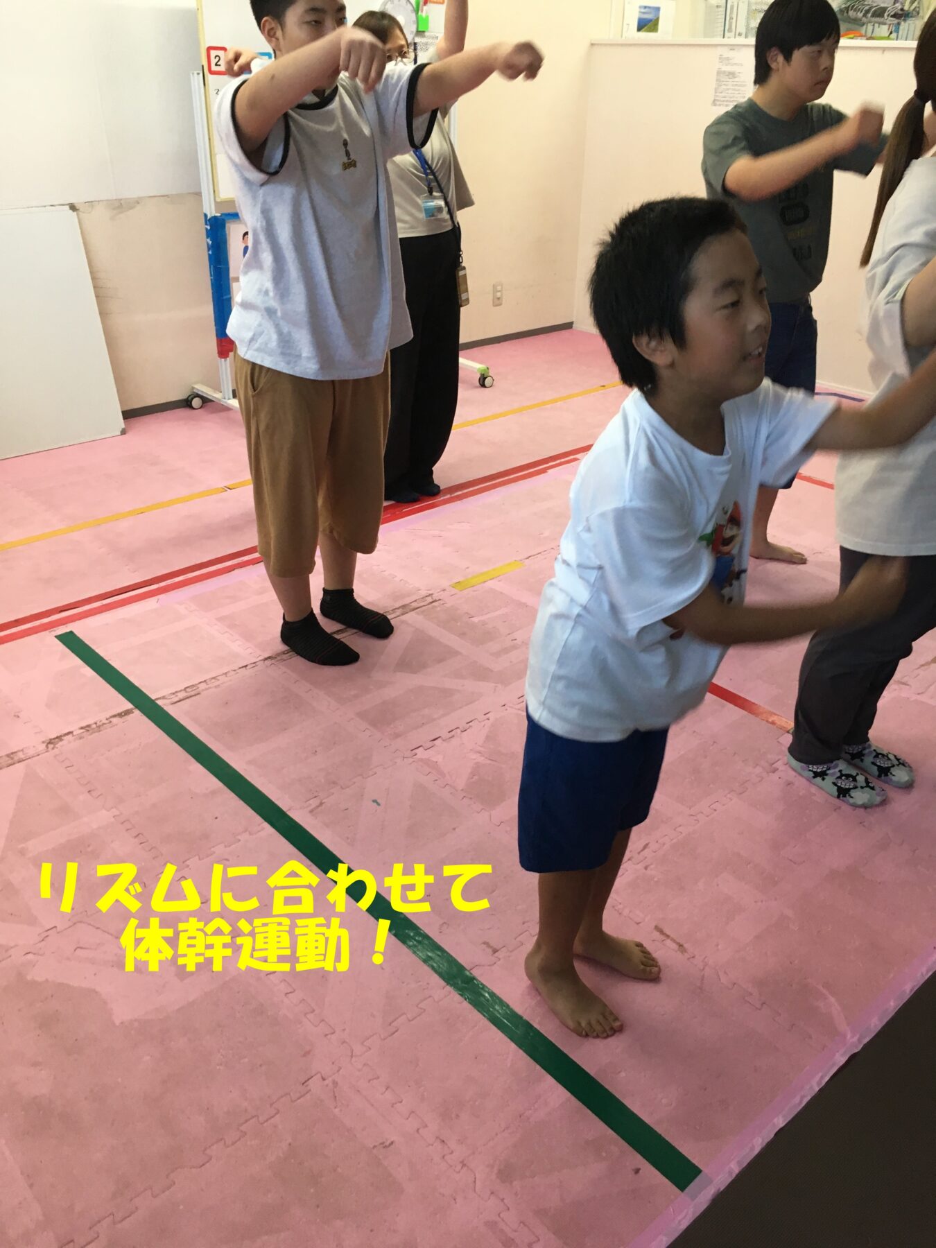Children in a gym, standing on pink mats, raising arms in a group exercise while a few observers watch in the background. A bright yellow caption in Japanese is visible at the bottom.