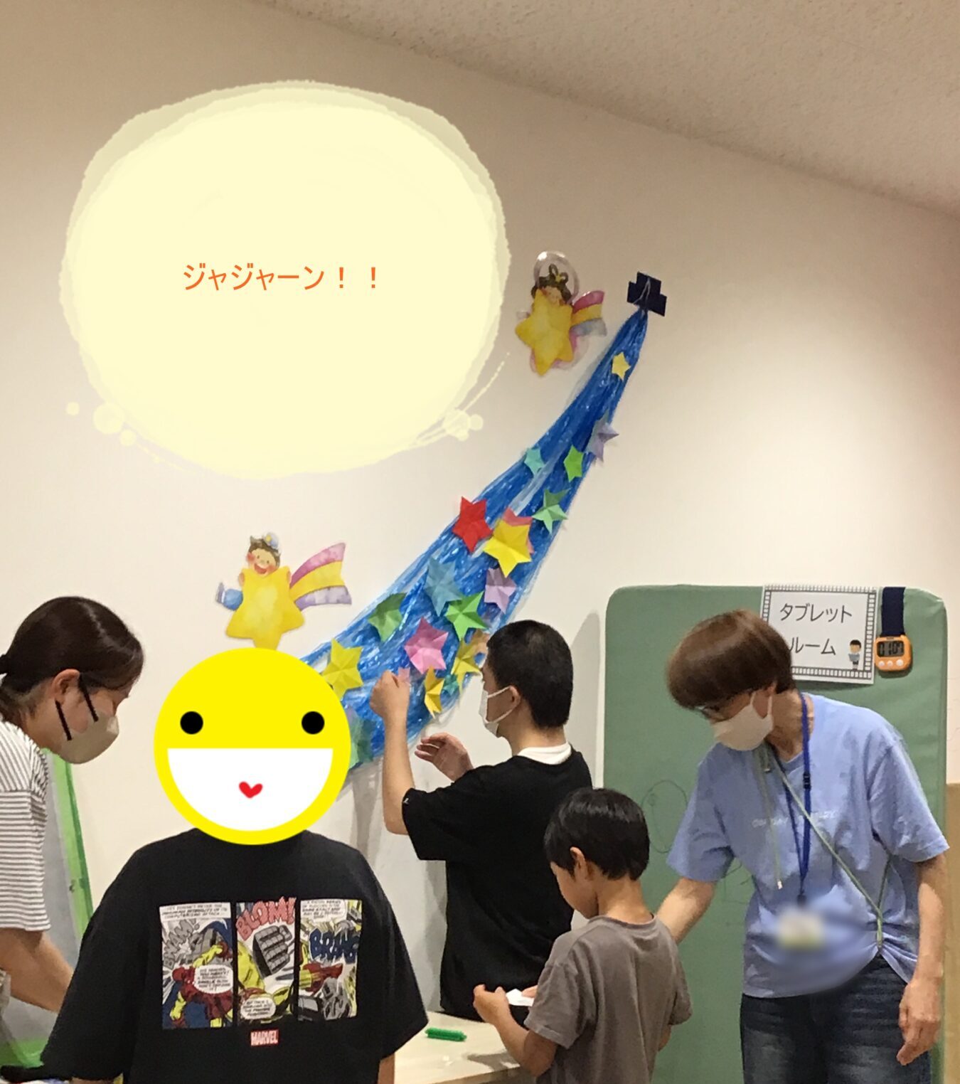 Group of people at a craft table under a wall mural with a yellow sun and a blue curved star trail; some wear masks.