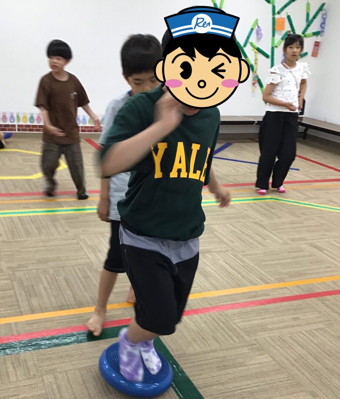 Child balances on a blue wobble disc in a gym while others watch, wearing a green Yale shirt and purple socks.