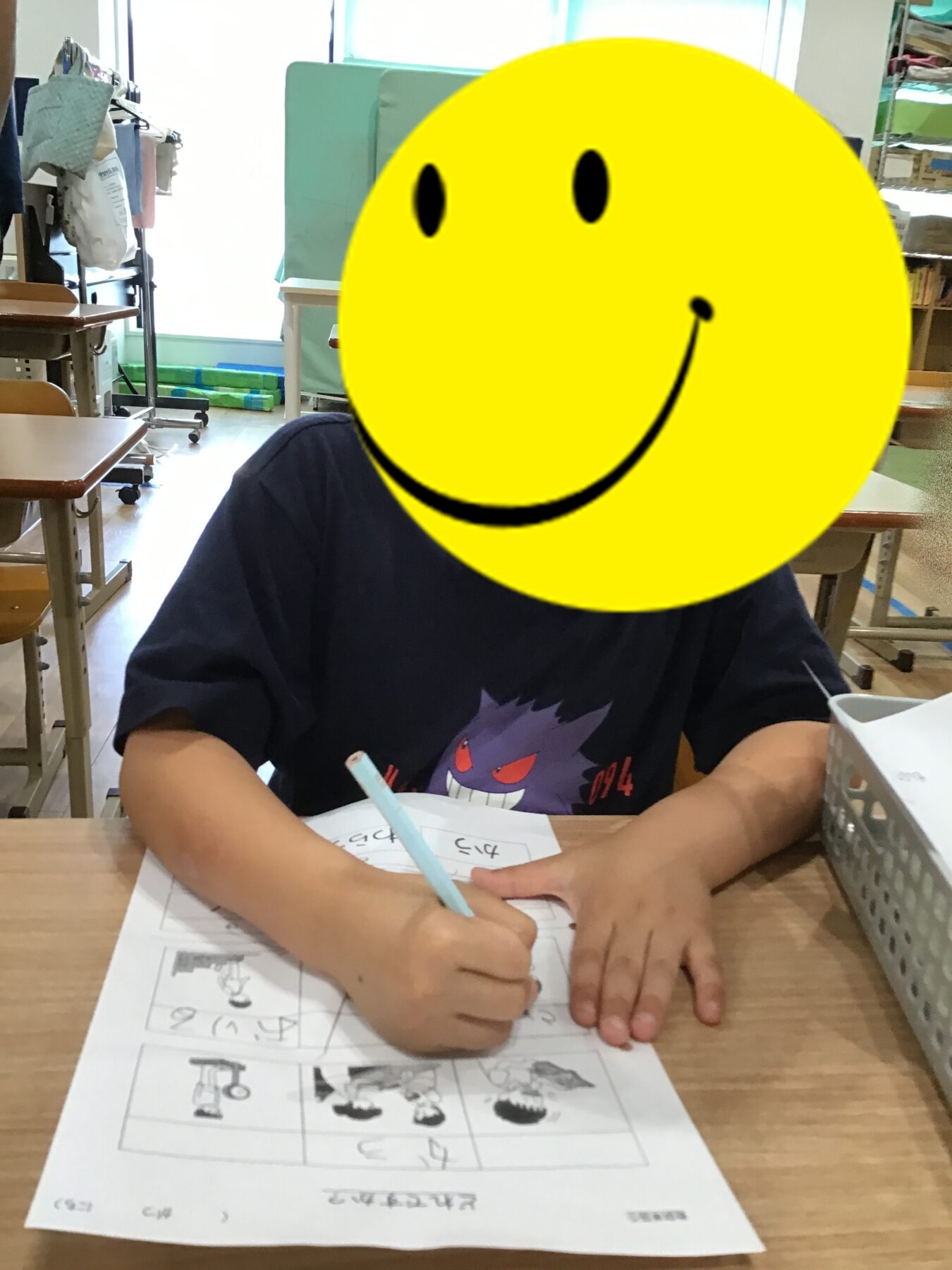 Child sitting at a desk in a classroom, writing on a worksheet; a large yellow smiley mask covers their face, obscuring identity.