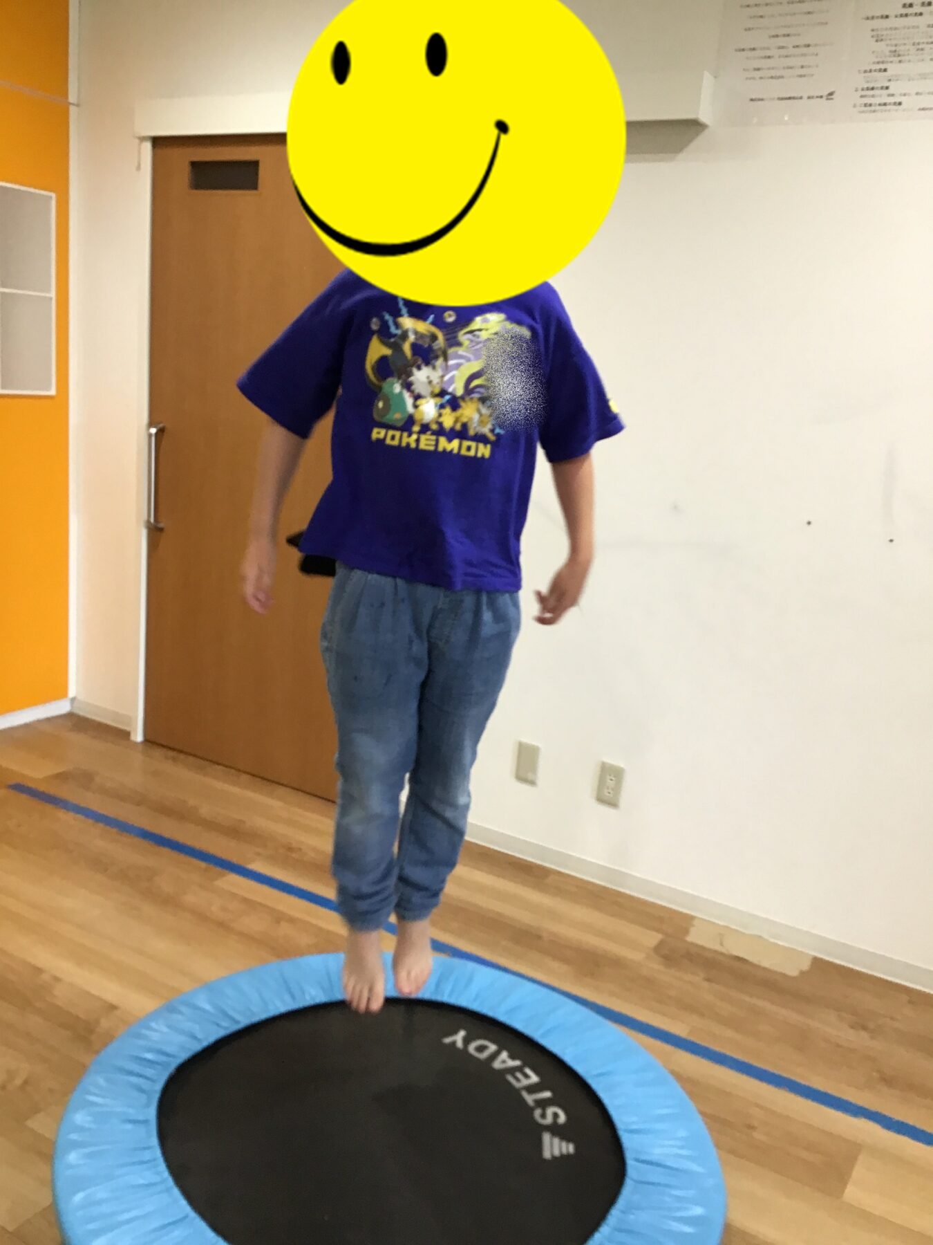 Child wearing a purple Pokémon shirt stands barefoot on a blue trampoline in an indoor room with orange wall and wooden floor, face covered by a large yellow smiley emoji.