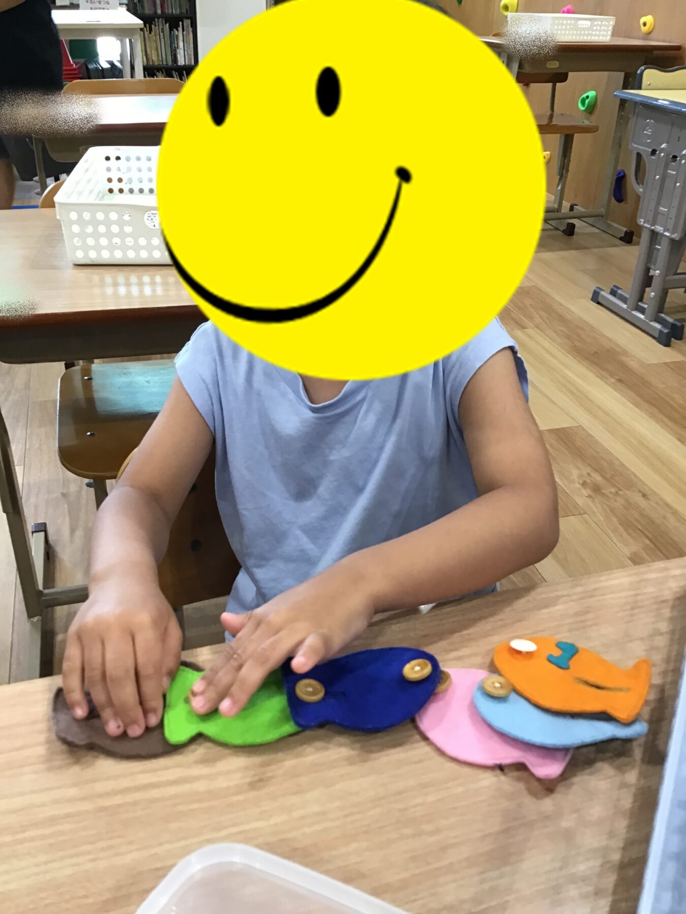 Child seated at a wooden table in a classroom, arranging colorful felt fish shapes with button eyes for a craft activity.