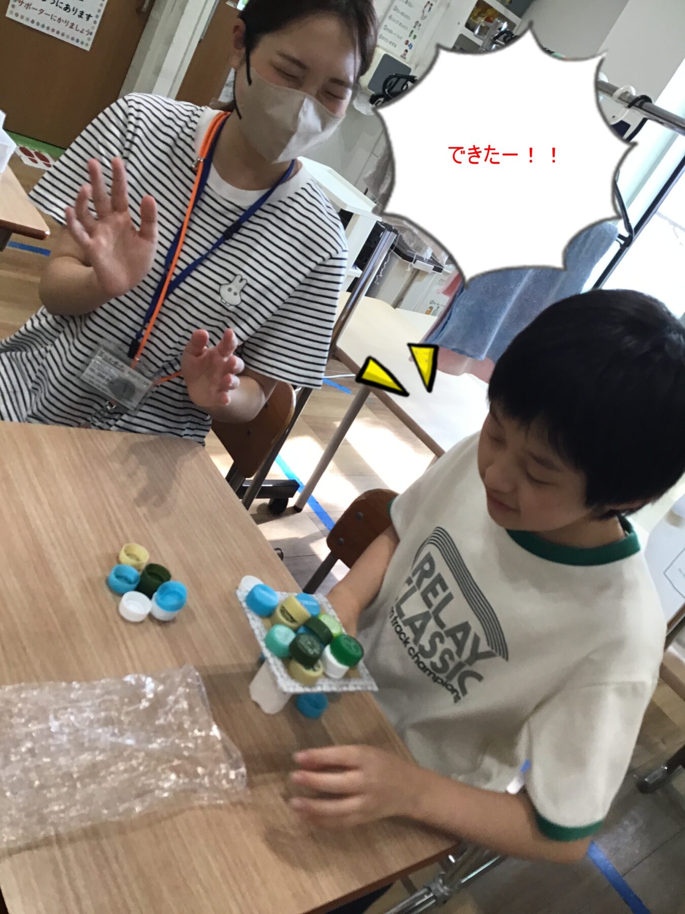 Caregiver in a striped shirt and mask sits with a child at a table during a painting activity, raising a hand in hello as paint pots lay nearby on the table close-up.