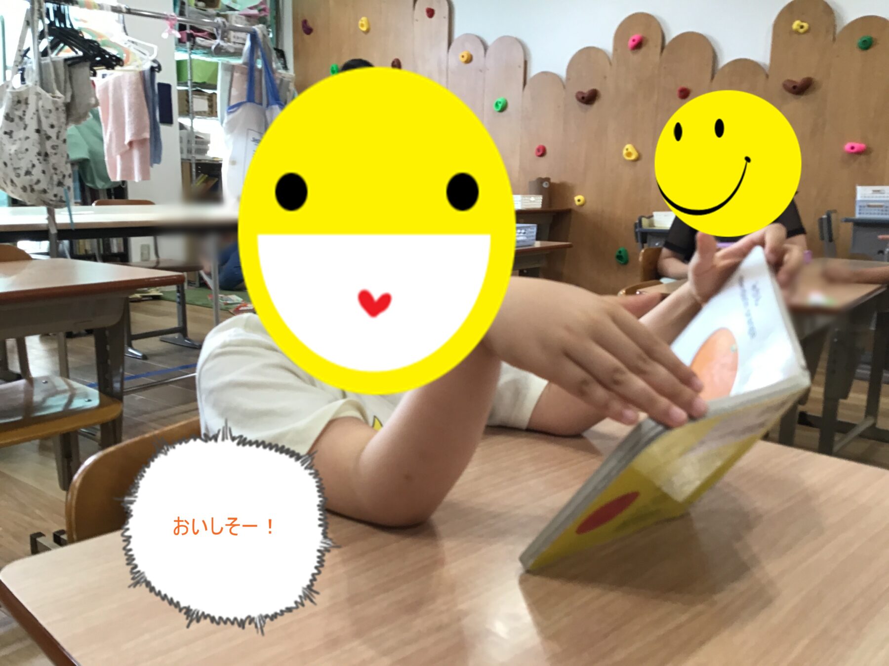 Two children sit at a classroom table reading a book; their faces are covered with large yellow smiley stickers. A speech bubble reads おいしそー!
