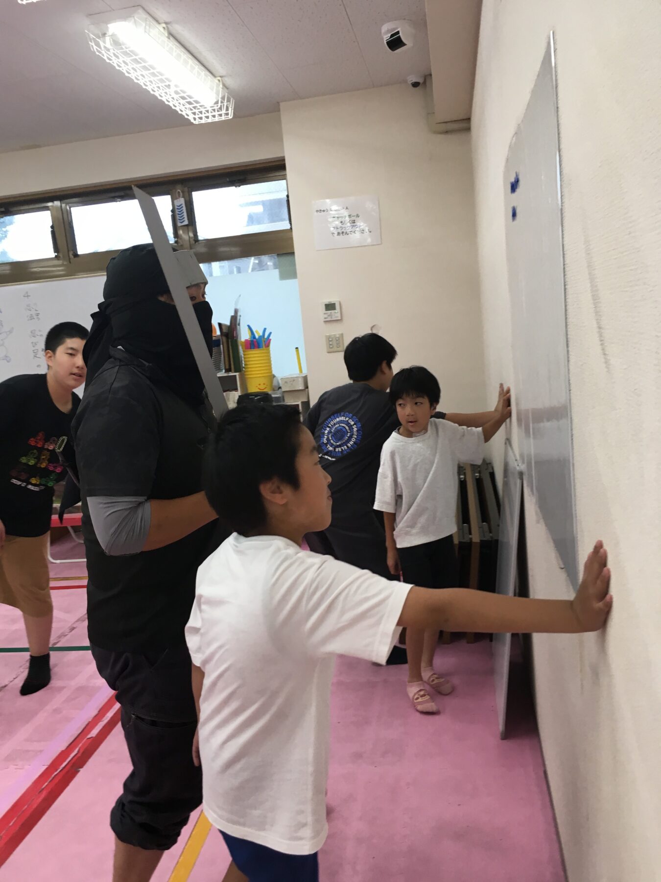 Instructor in black athletic gear with a sword prop watches over children as they lean against a wall in a gym activity, on pink mats.