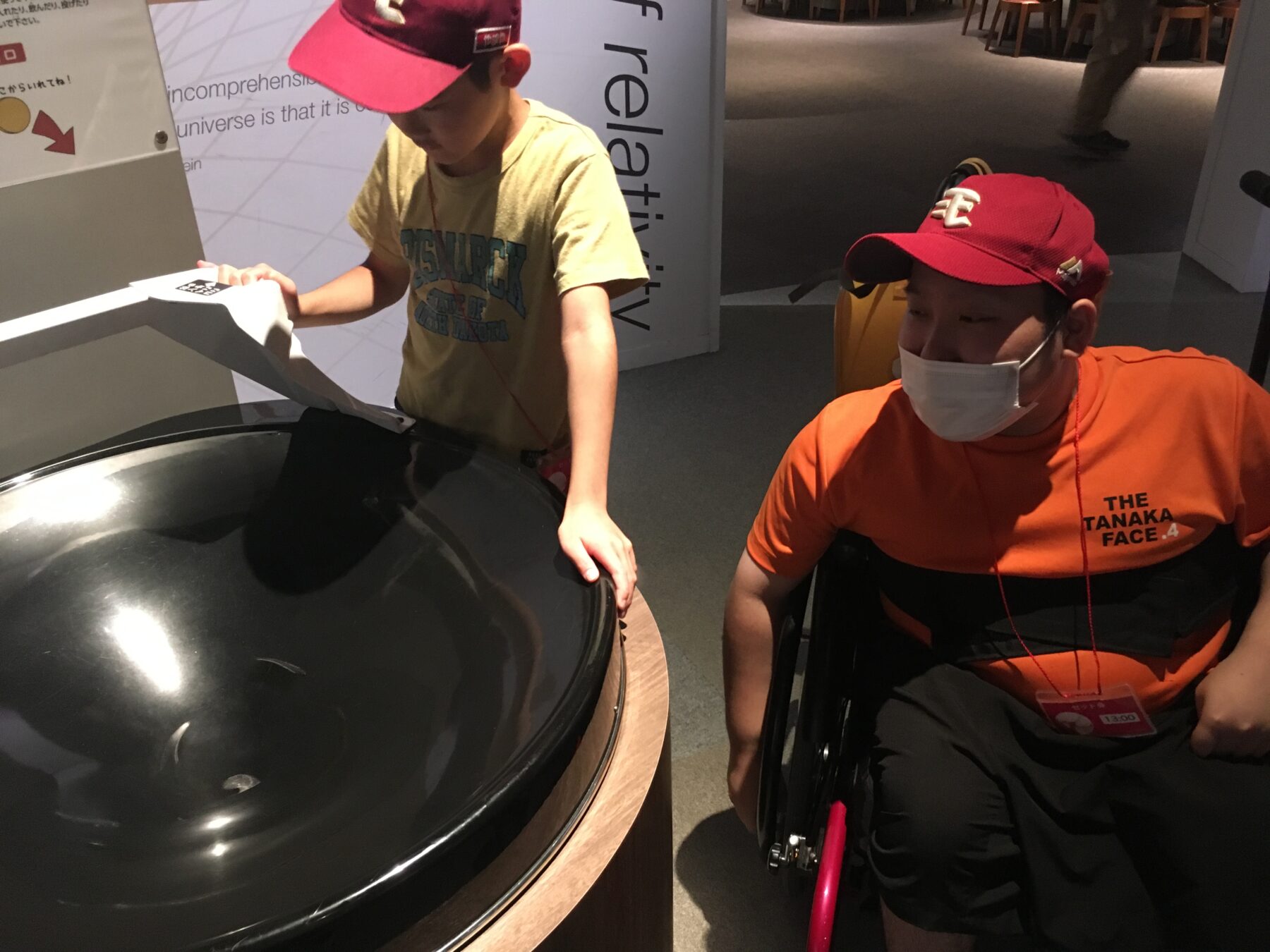 Two people—one boy in a yellow shirt and pink cap placing a white sheet onto a large black circular exhibit, while a person in a bright orange shirt and red cap sits in a wheelchair nearby, wearing a mask.