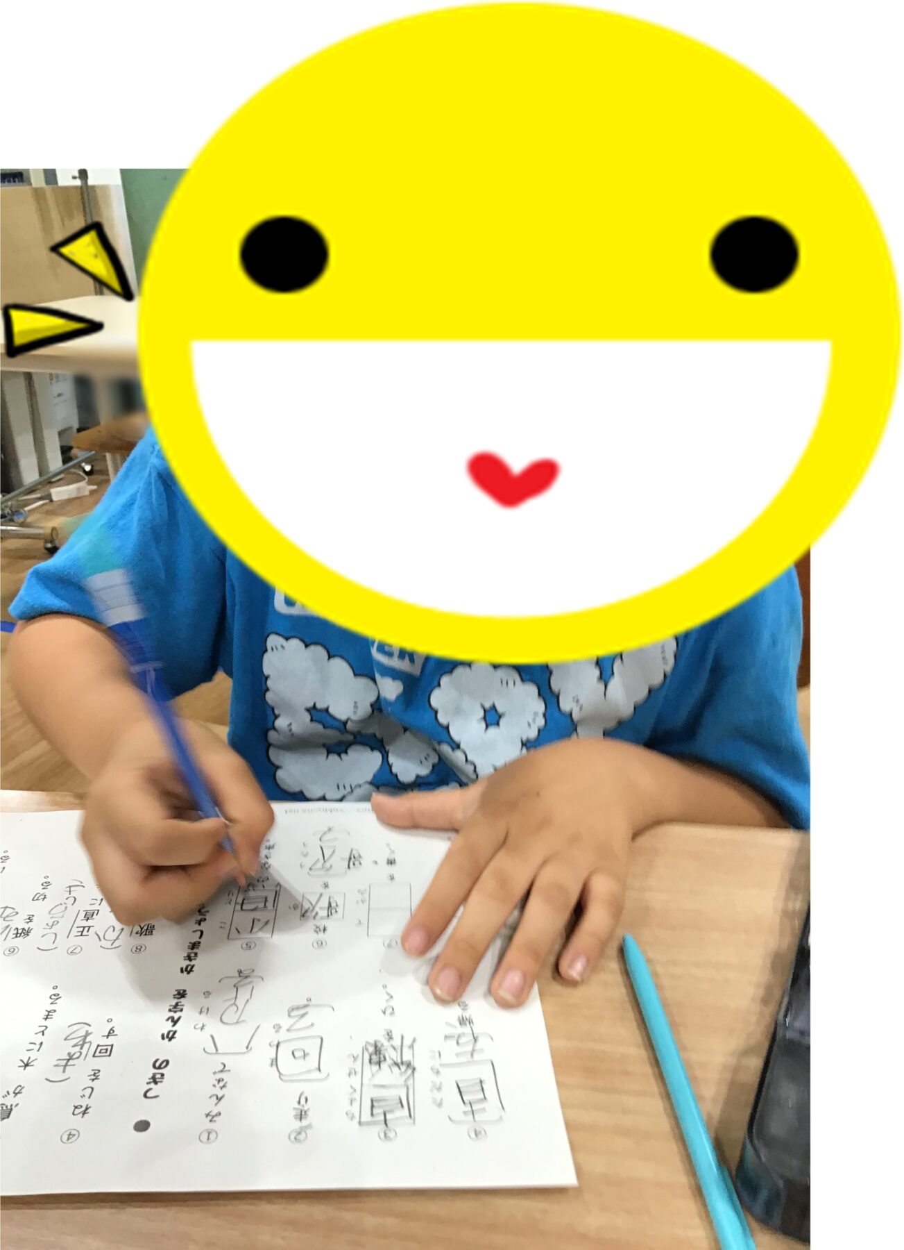 Child seated at a desk writing on a worksheet, with a large yellow smiley-face sticker covering their face.