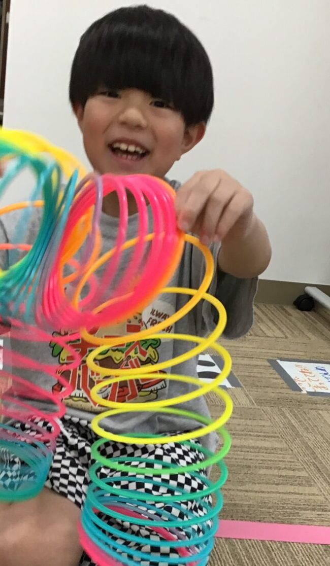 Smiling child sits on the floor playing with colorful spiral coil toys stacked in his hands and lap.
