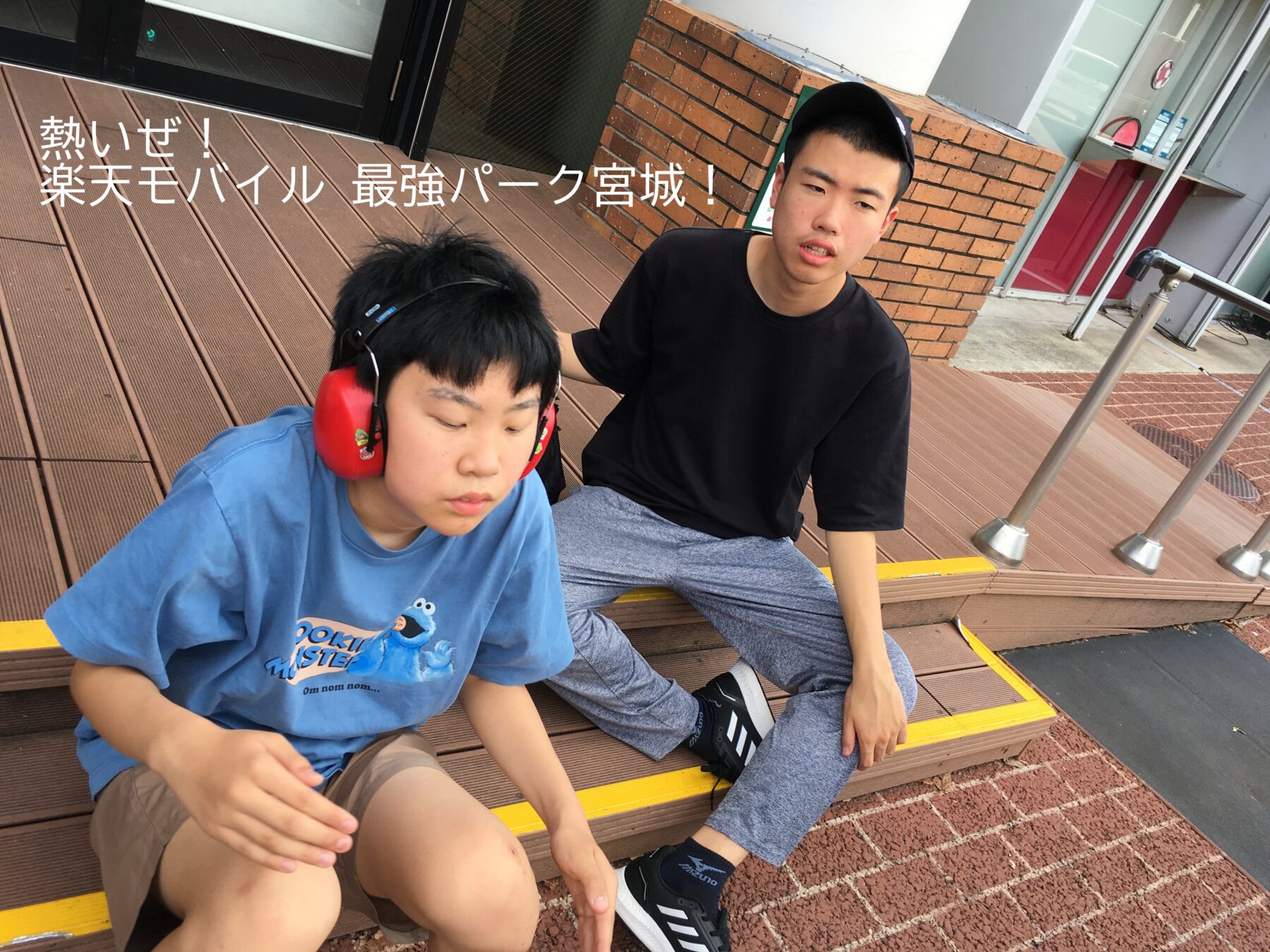 Two young men rest on brown outdoor steps by a brick wall and glass storefront; one wears red hearing protection and a Cookie Monster blue shirt, the other a black t‑shirt and cap.