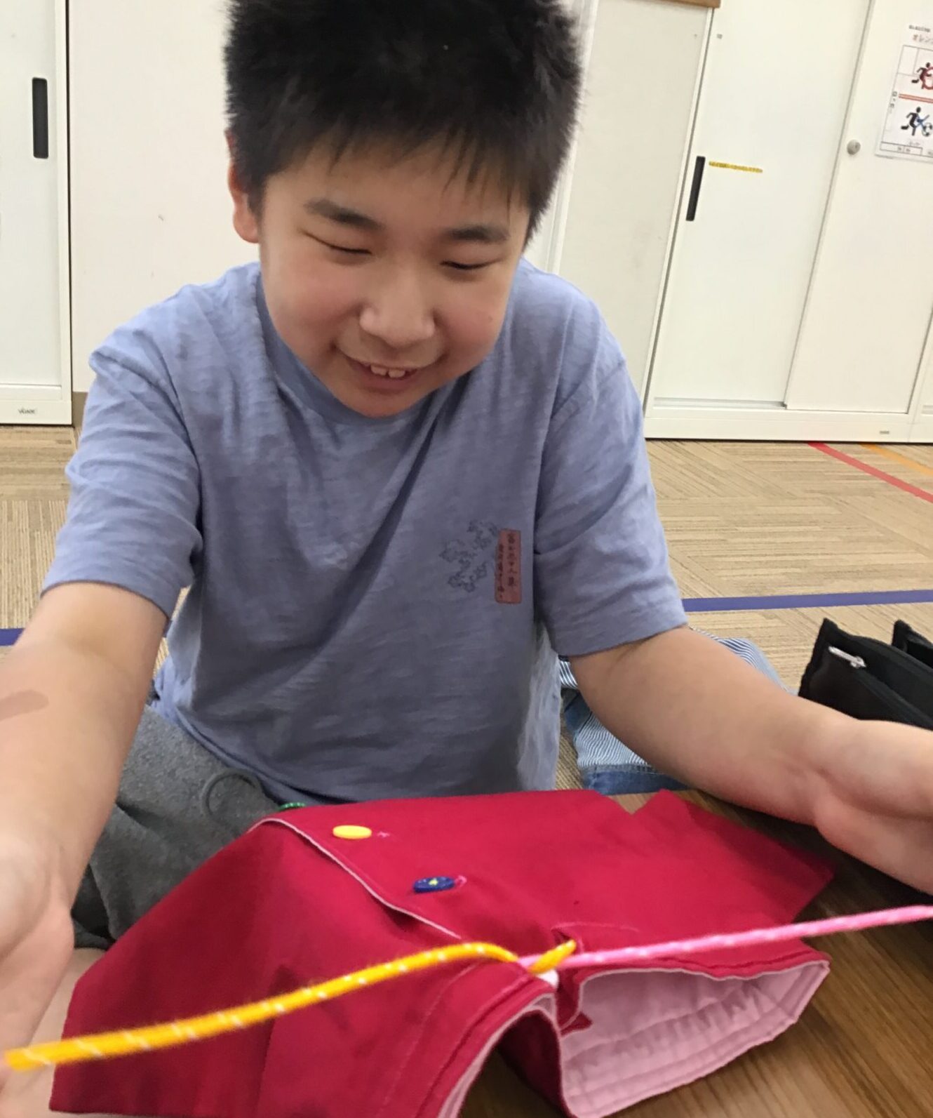 Boy or student smiling while sewing bright red fabric with yellow piping and colorful buttons in a craft room or classroom setting.